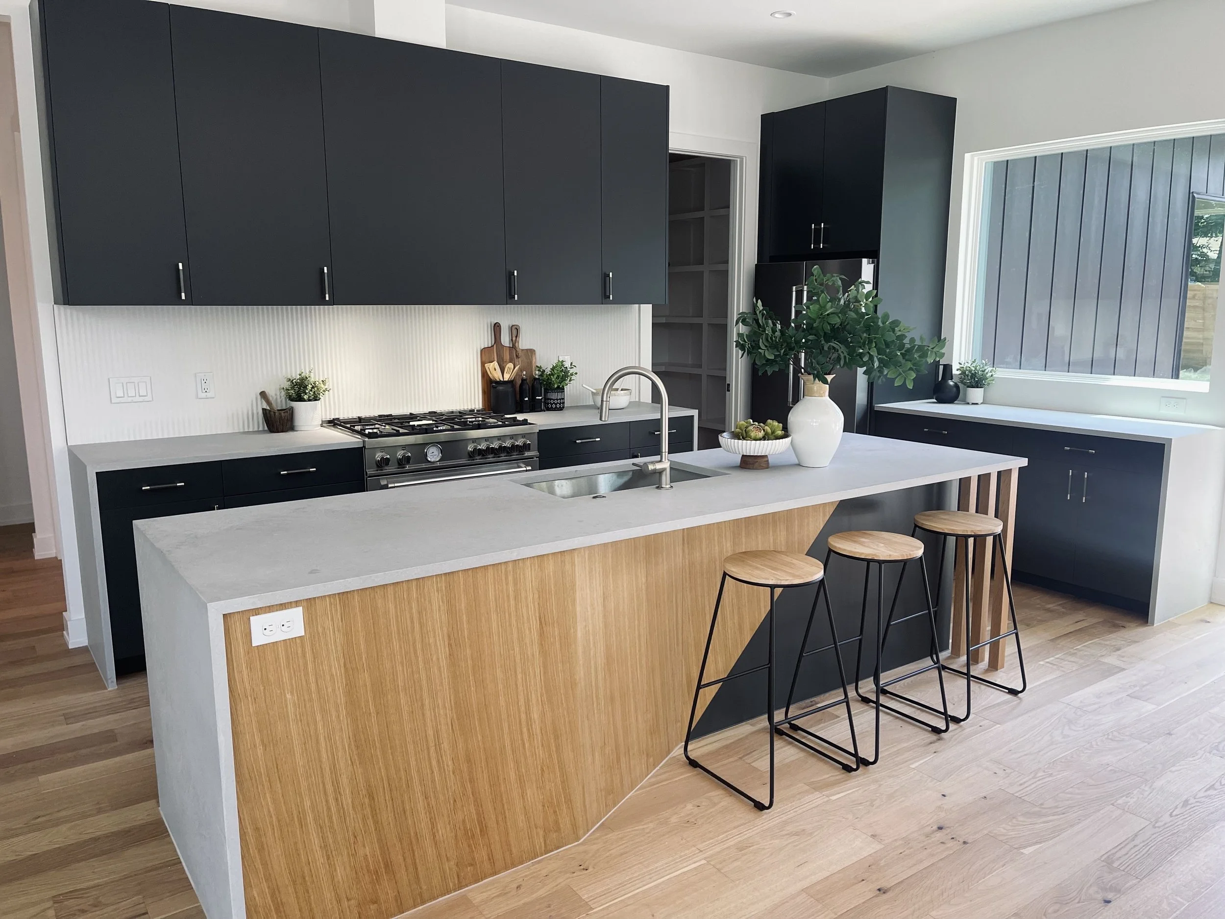 Modern kitchen with dark cabinetry, a white kitchen island with a wooden panel, three wooden bar stools, a window with a view outside, and decorative plants.