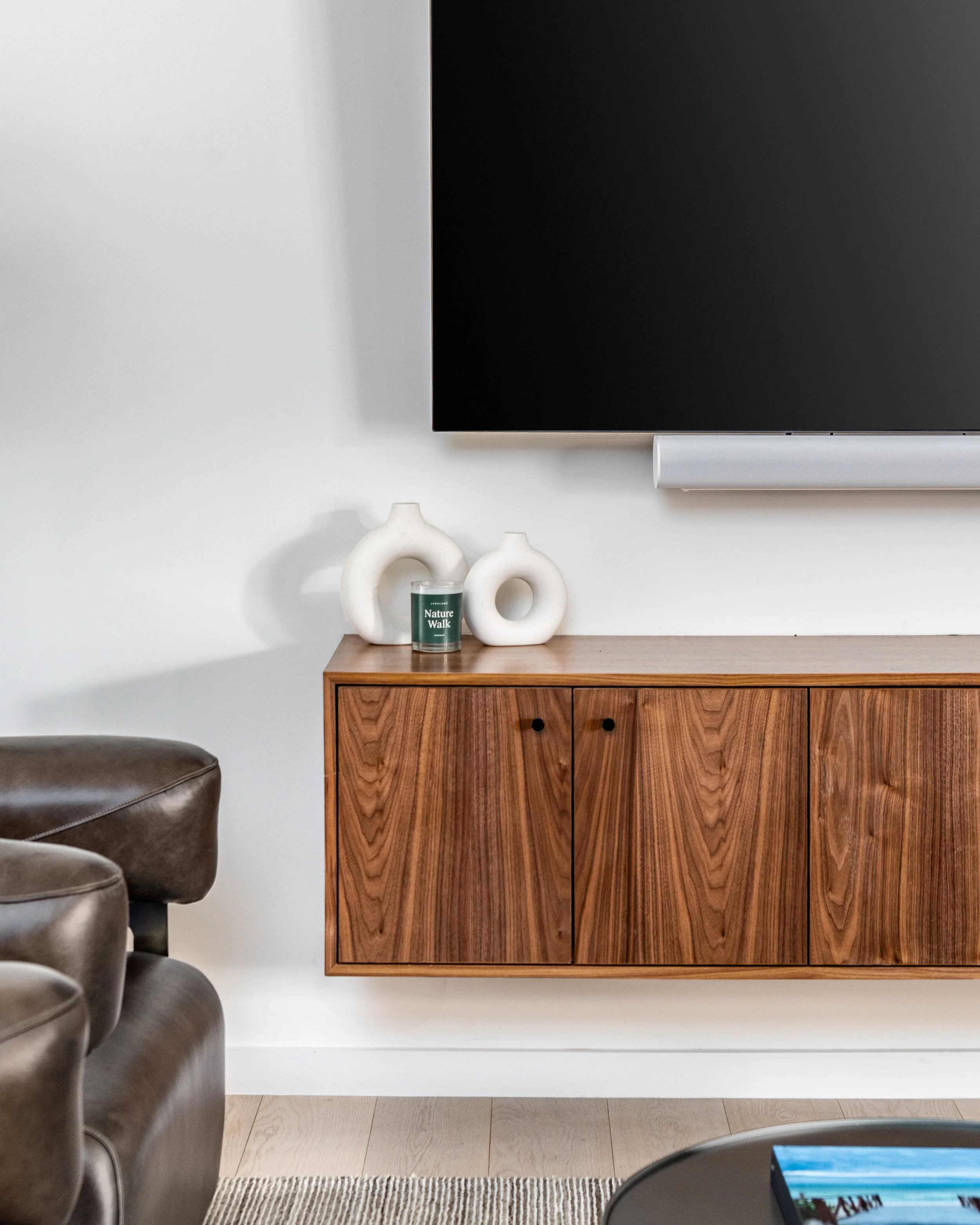 Close-up of a modern living room corner showing a wooden wall-mounted cabinet with white vases and a candle, a large flat-screen TV, and part of a leather sofa.