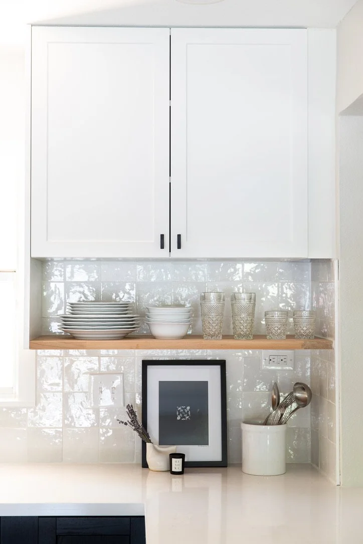 White kitchen upper cabinet with black handles, open shelf with stacked white and gray plates, clear textured glasses, framed artwork, and kitchen utensils on a white countertop.