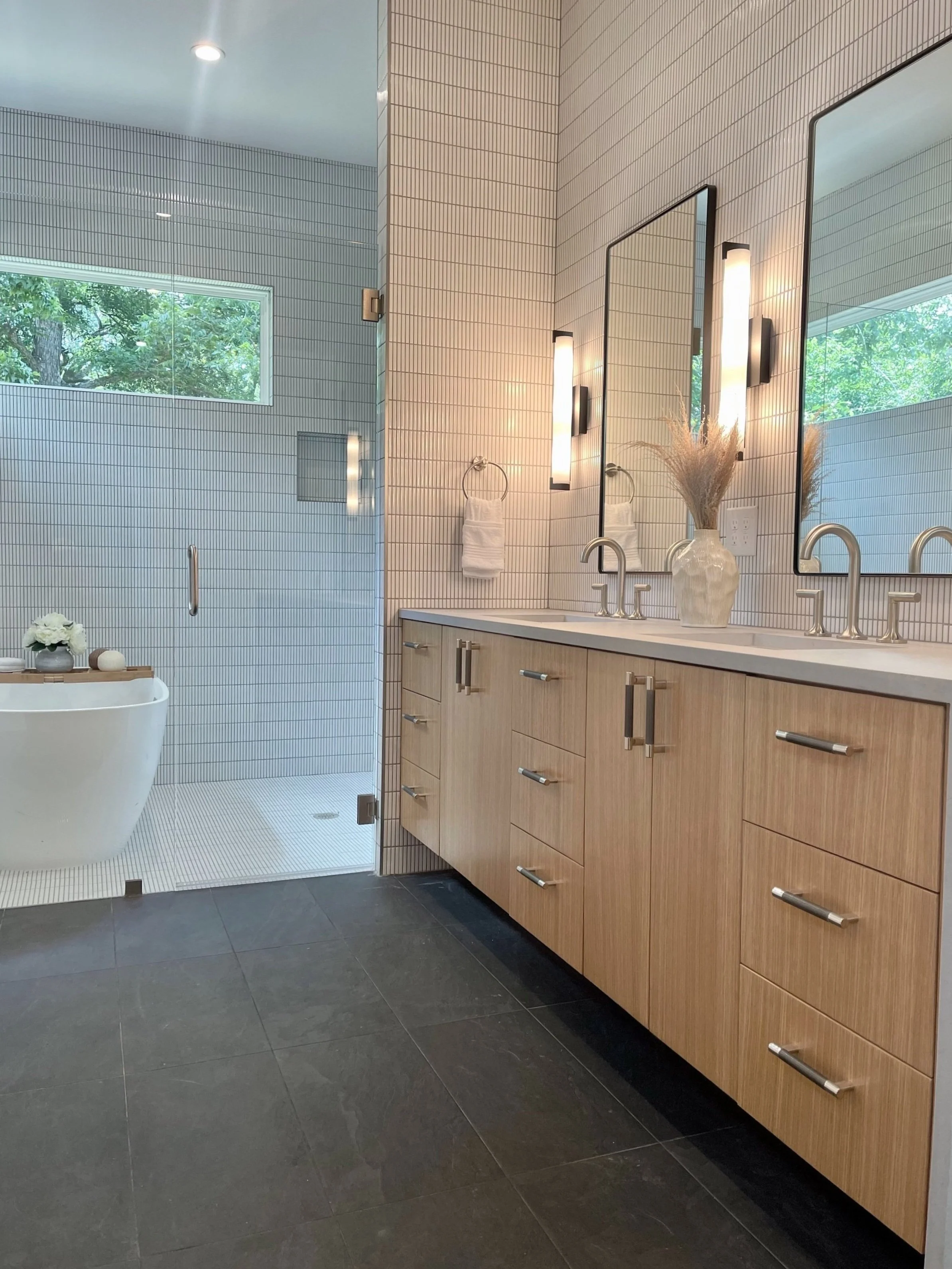 Modern bathroom with dual sinks, wooden cabinetry, large mirrors, wall-mounted lights, decorative vase with pampas grass, and a walk-in shower with glass door, white tiled walls, and a window with greenery outside.