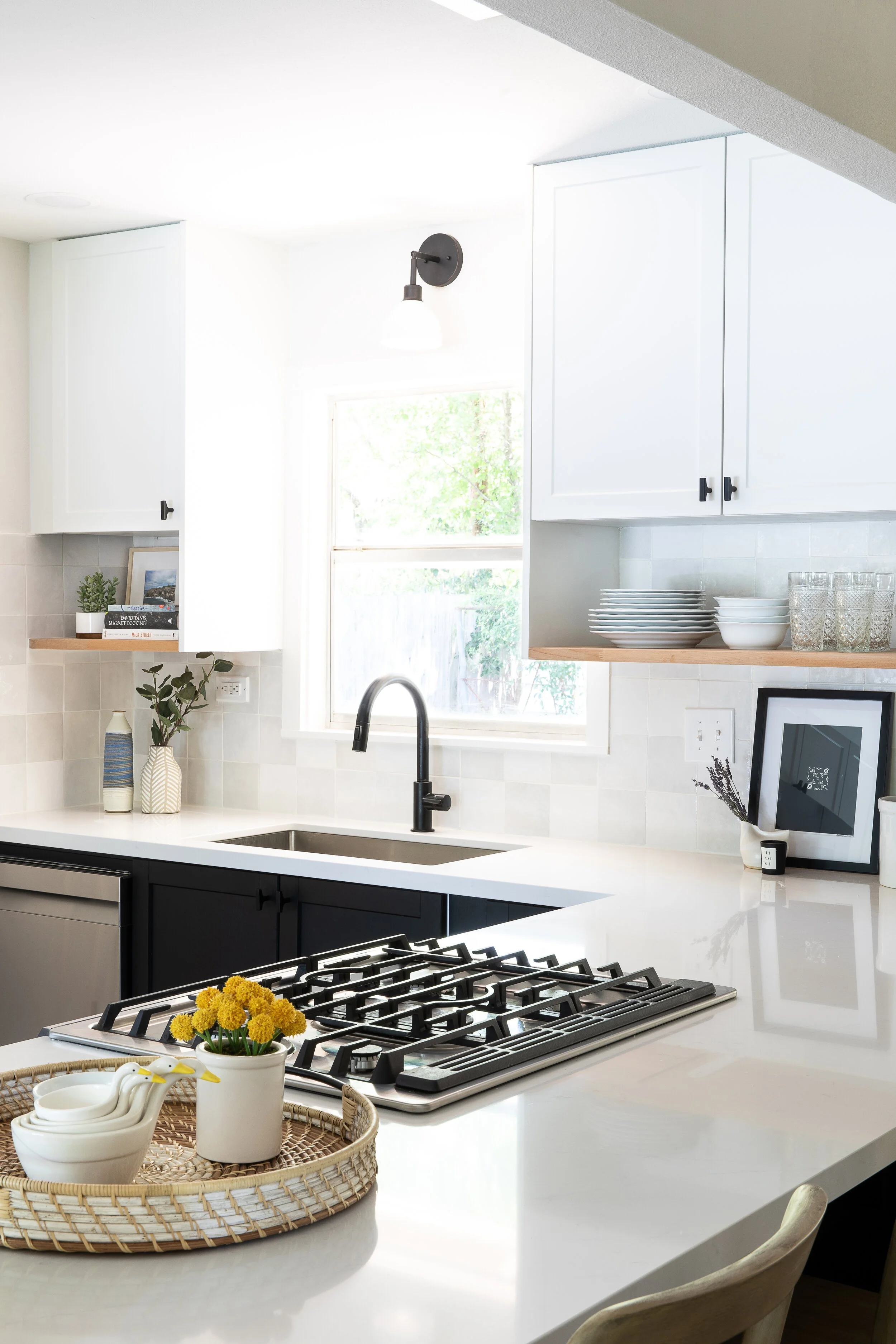 Modern kitchen with white cabinets, black hardware, and a white countertop island. On the island, there's a wicker tray with white ceramic bowls and a small potted yellow flowering plant. The backsplash features white tiles, and there's a black fauce