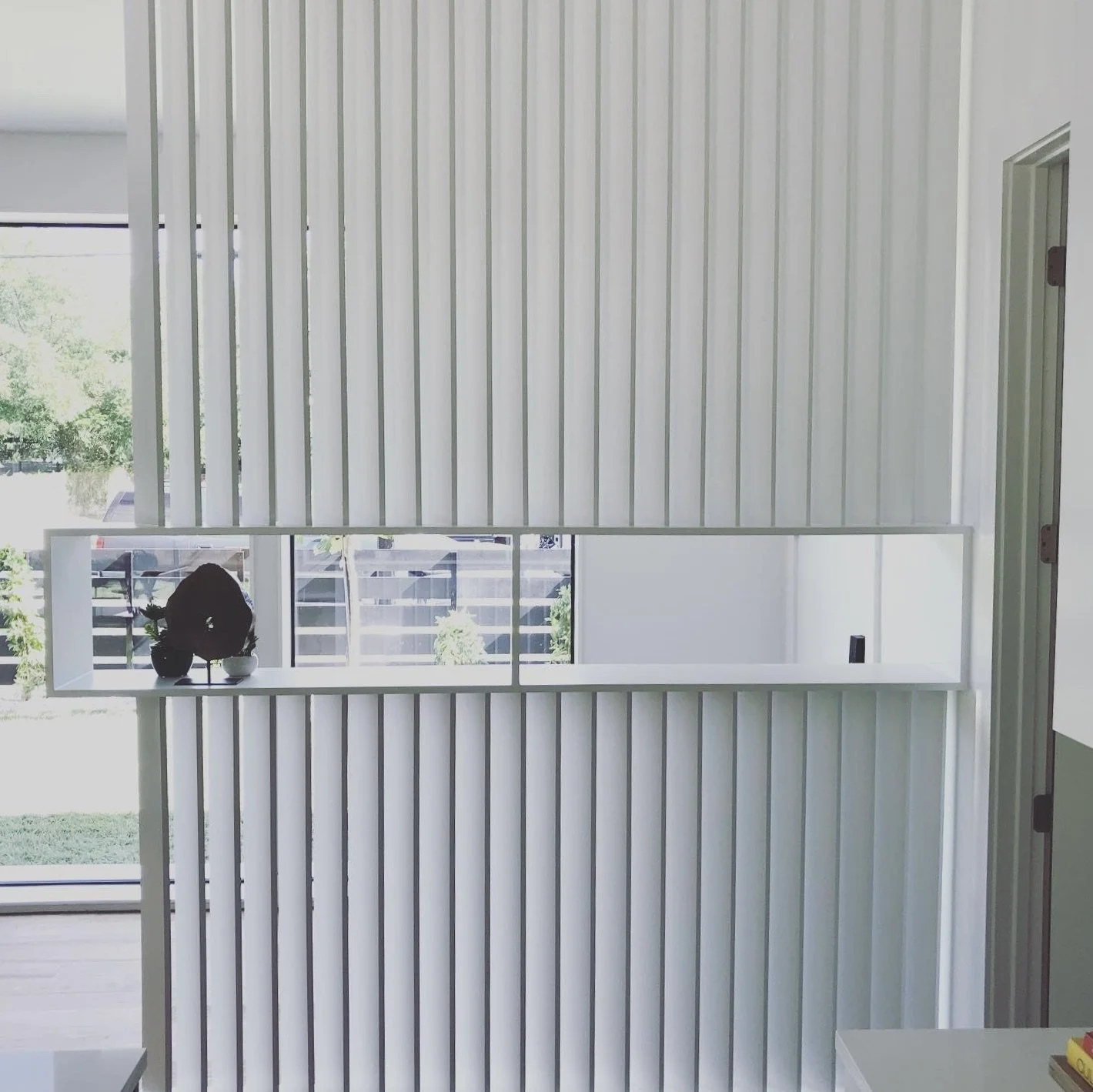 Interior view of a modern room with white vertical paneling and a large rectangular mirror reflecting a window and outdoor scenery, with a small black vase and plant on a ledge in front of the mirror.