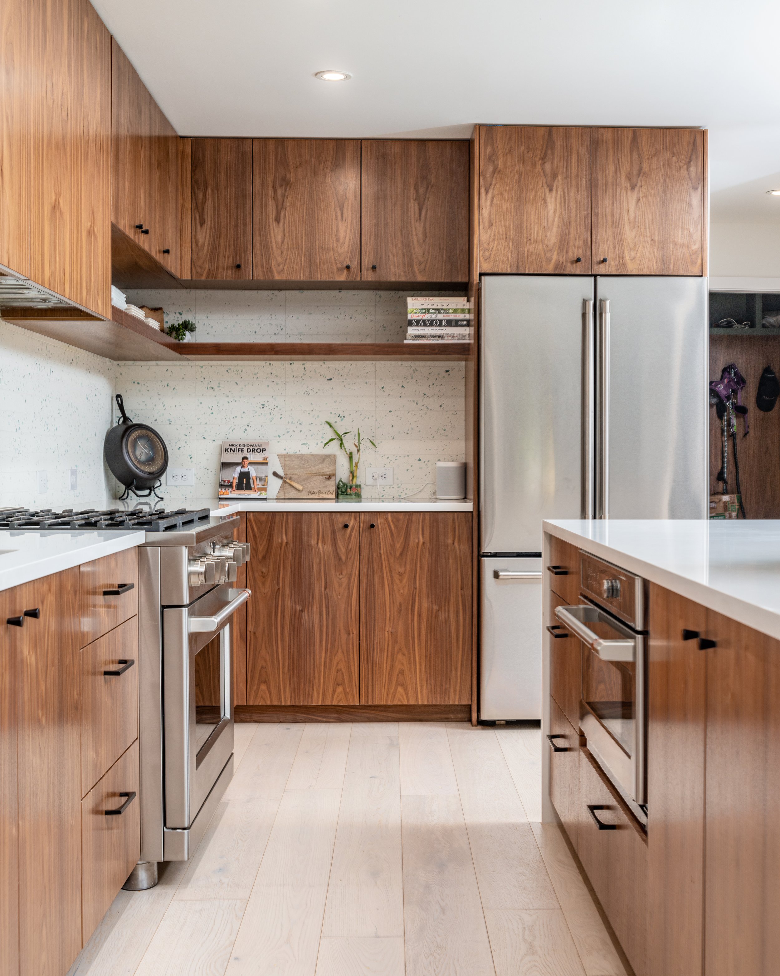 Modern kitchen with wooden cabinets, stainless steel appliances, a white countertop, and minimal decor including books, plants, and a cookbook, with light-colored flooring.