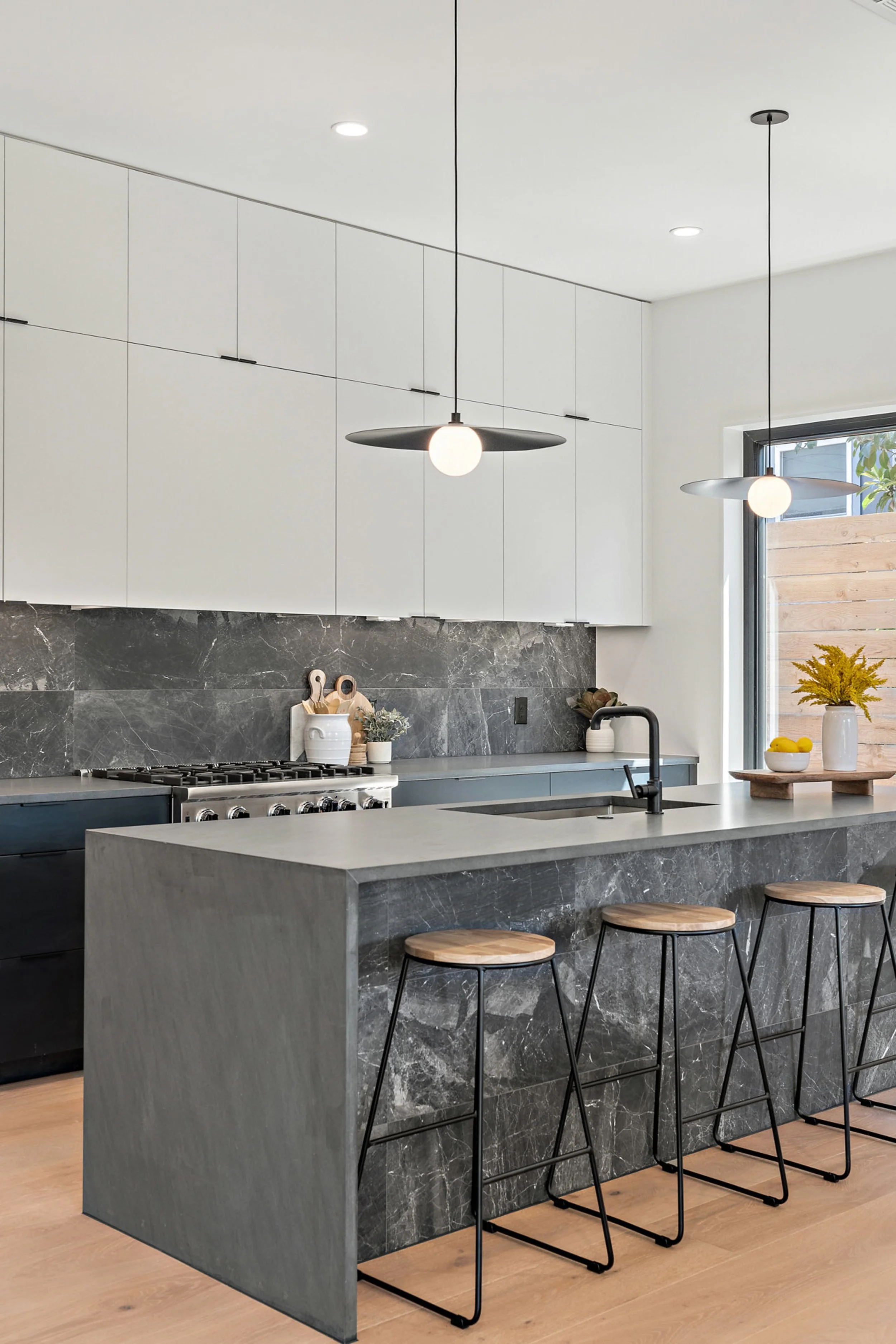 Modern kitchen with white upper cabinets, black and gray marble backsplash, gray marble kitchen island, black faucet, three wooden barstools with black metal legs, pendant lights, window with a view of a wooden fence and plants, decorative bowls with