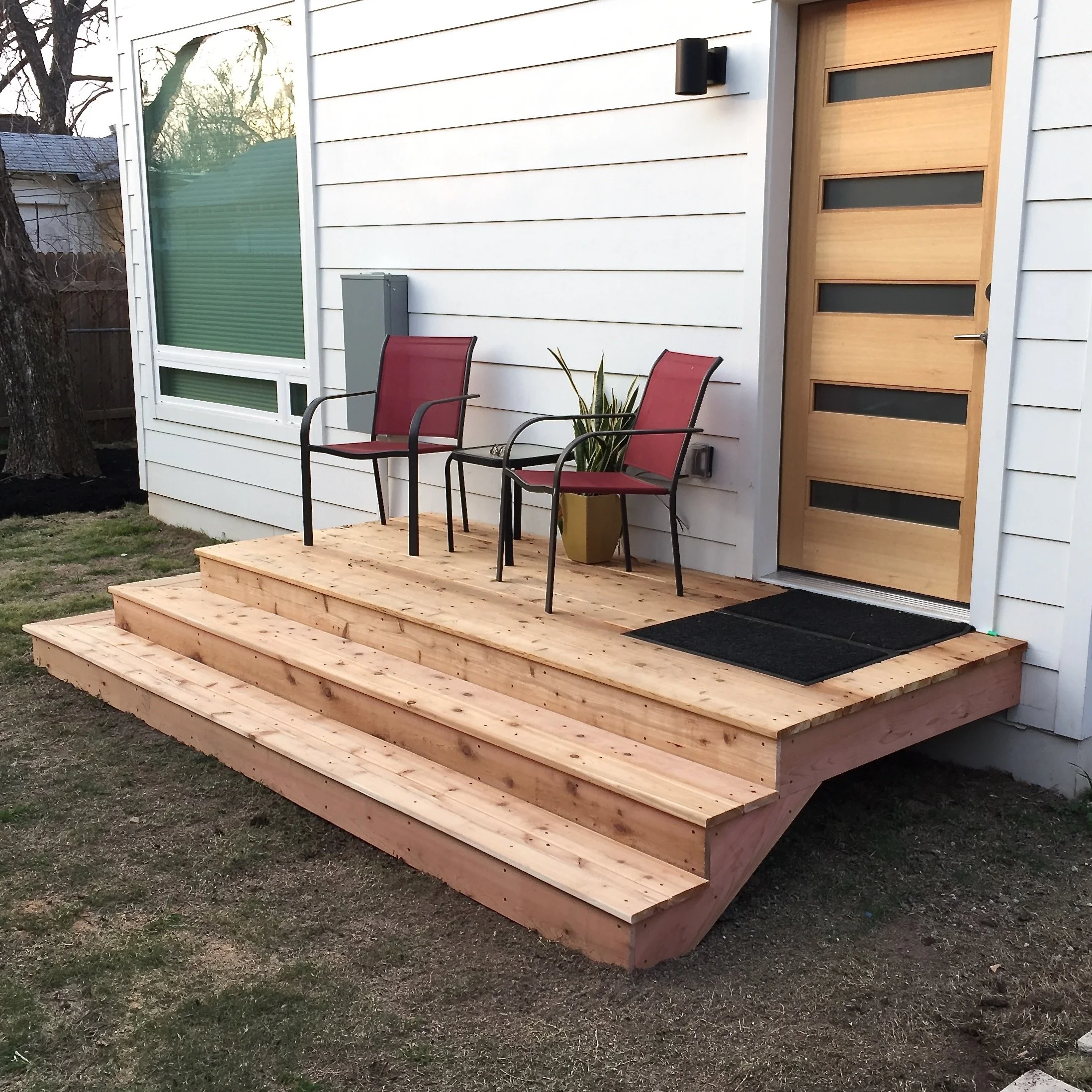 Wooden front porch with two red chairs, a potted plant, and a black doormat in front of a modern wooden door with horizontal glass panels.