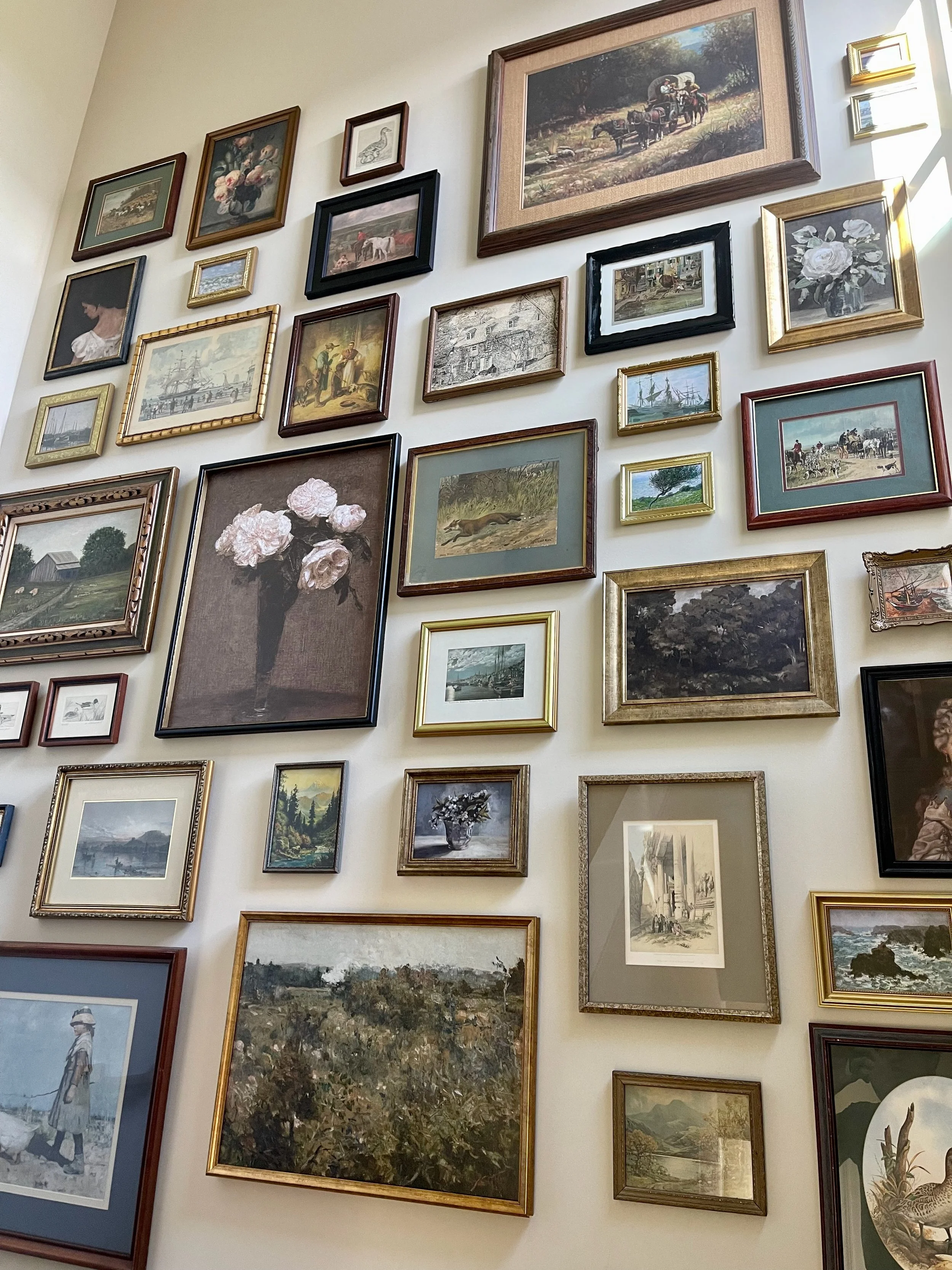 A gallery wall with various framed paintings and photographs, including landscapes, animals, flowers, and people.