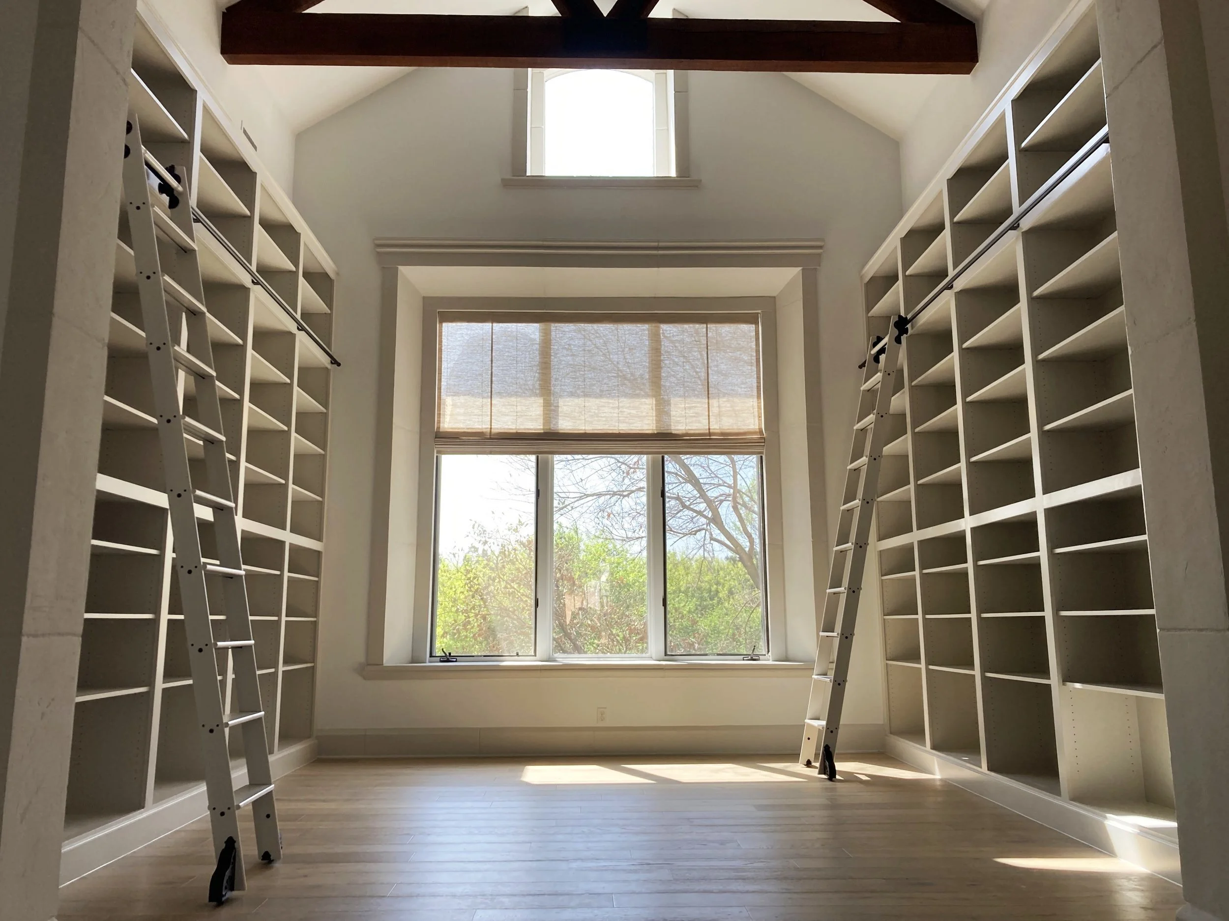 A bright room with large windows, built-in white shelves on both sides, and two ladders leaning against the shelves.