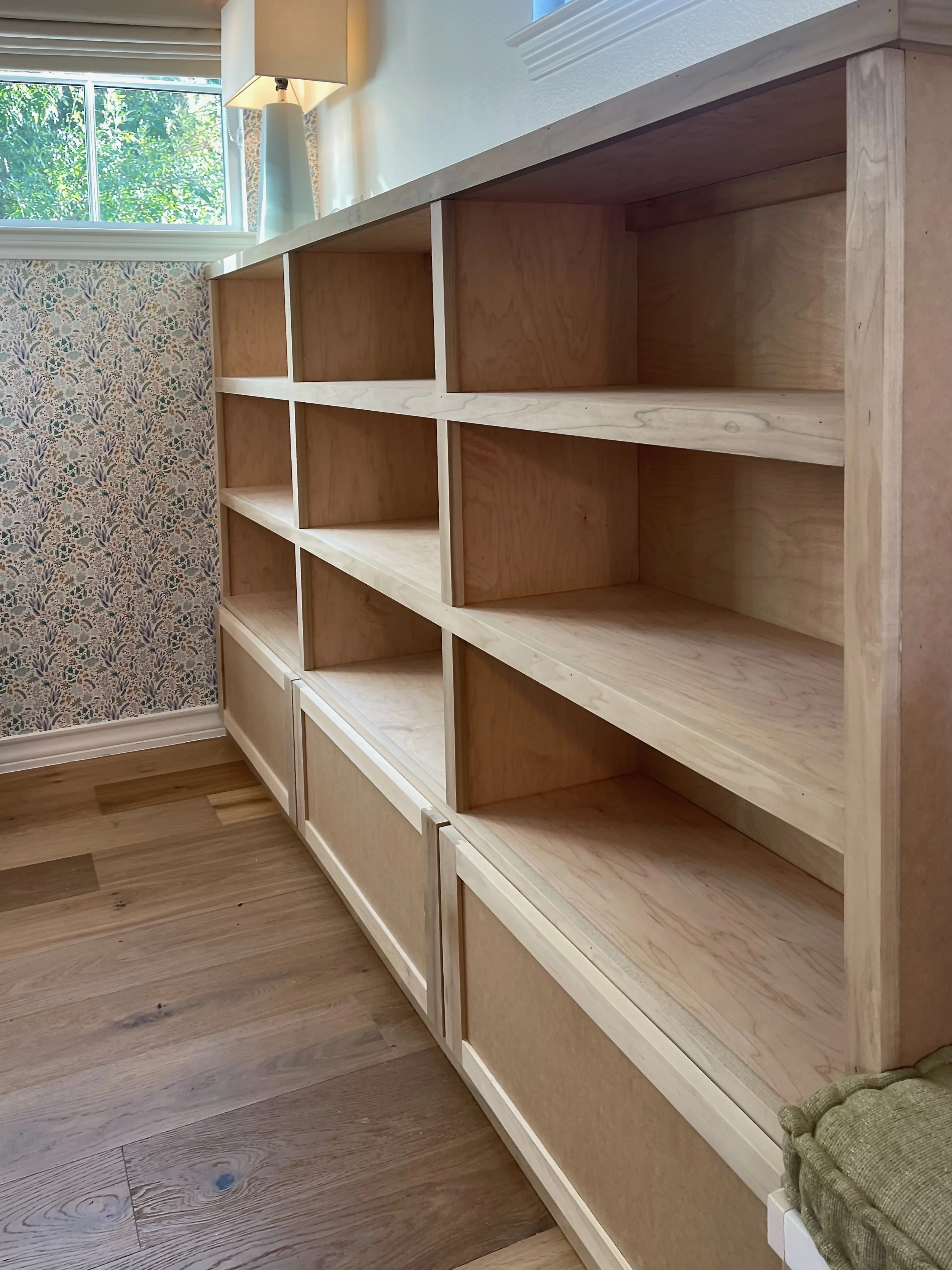 Empty wooden bookshelf with multiple shelves in a room with a window, a patterned wall, a lamp, and hardwood flooring.