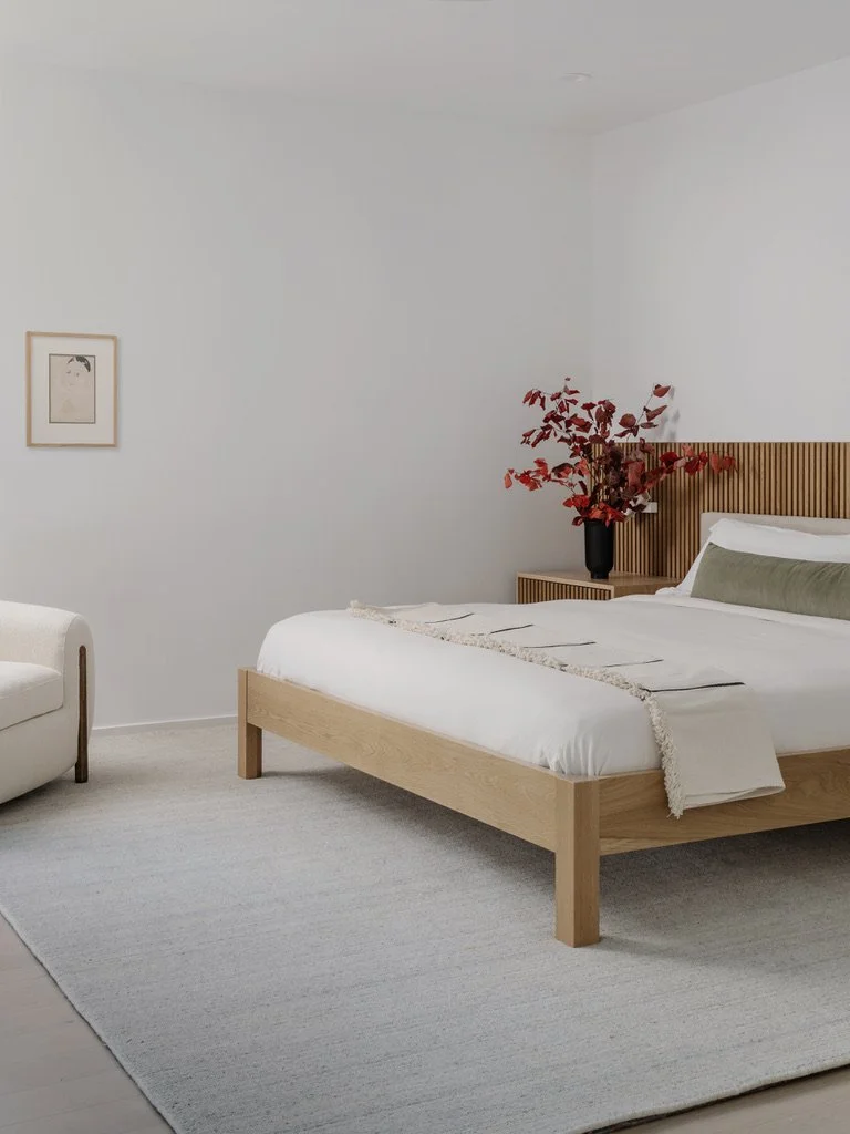 A minimalistic bedroom with a wooden bedframe, white bedding, a green pillow, a small side table with a flower vase, a framed artwork on the wall, and a white armchair, all against white walls and a light-colored rug.