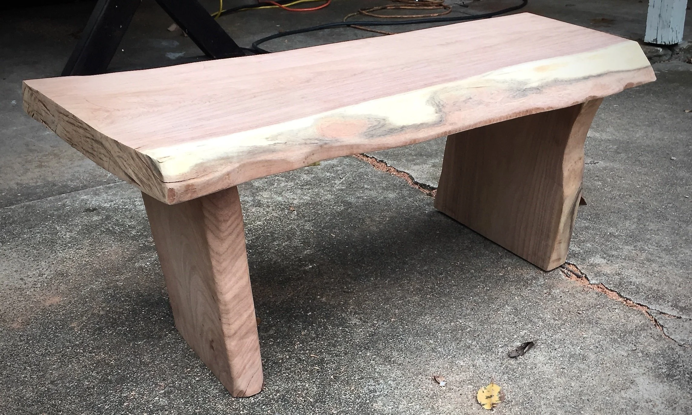 Wooden bench with a thick, natural edge seat and two rectangular legs, placed on a concrete surface outside.