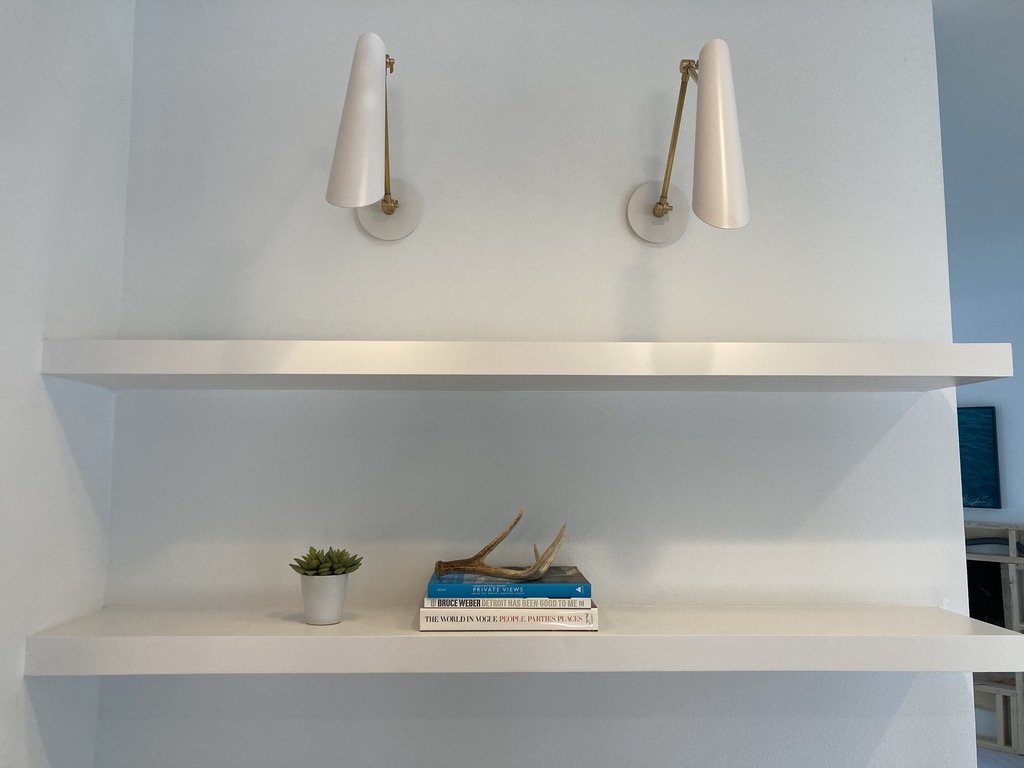 Two white adjustable wall-mounted lamps above two white floating shelves, with a potted succulent, a small decorative animal skull, and three books on the lower shelf.