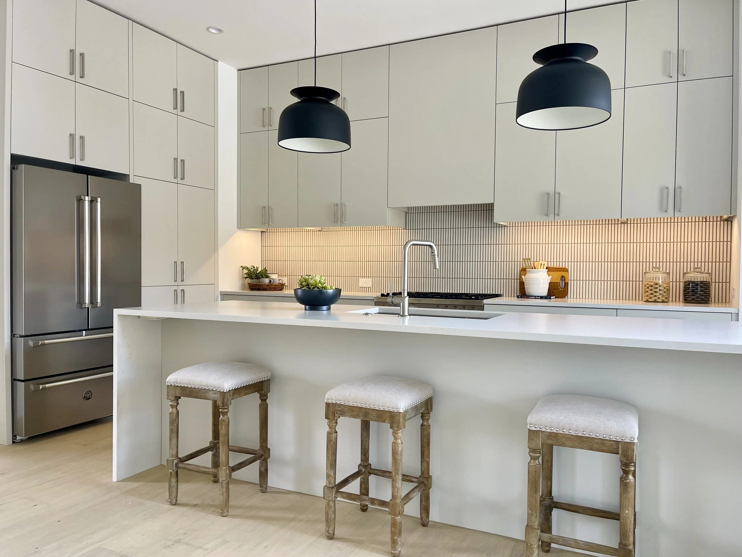 Modern kitchen with white cabinets, stainless steel refrigerator, black hanging pendant lights, beige bar stools, and a white island with a bowl of green apples.