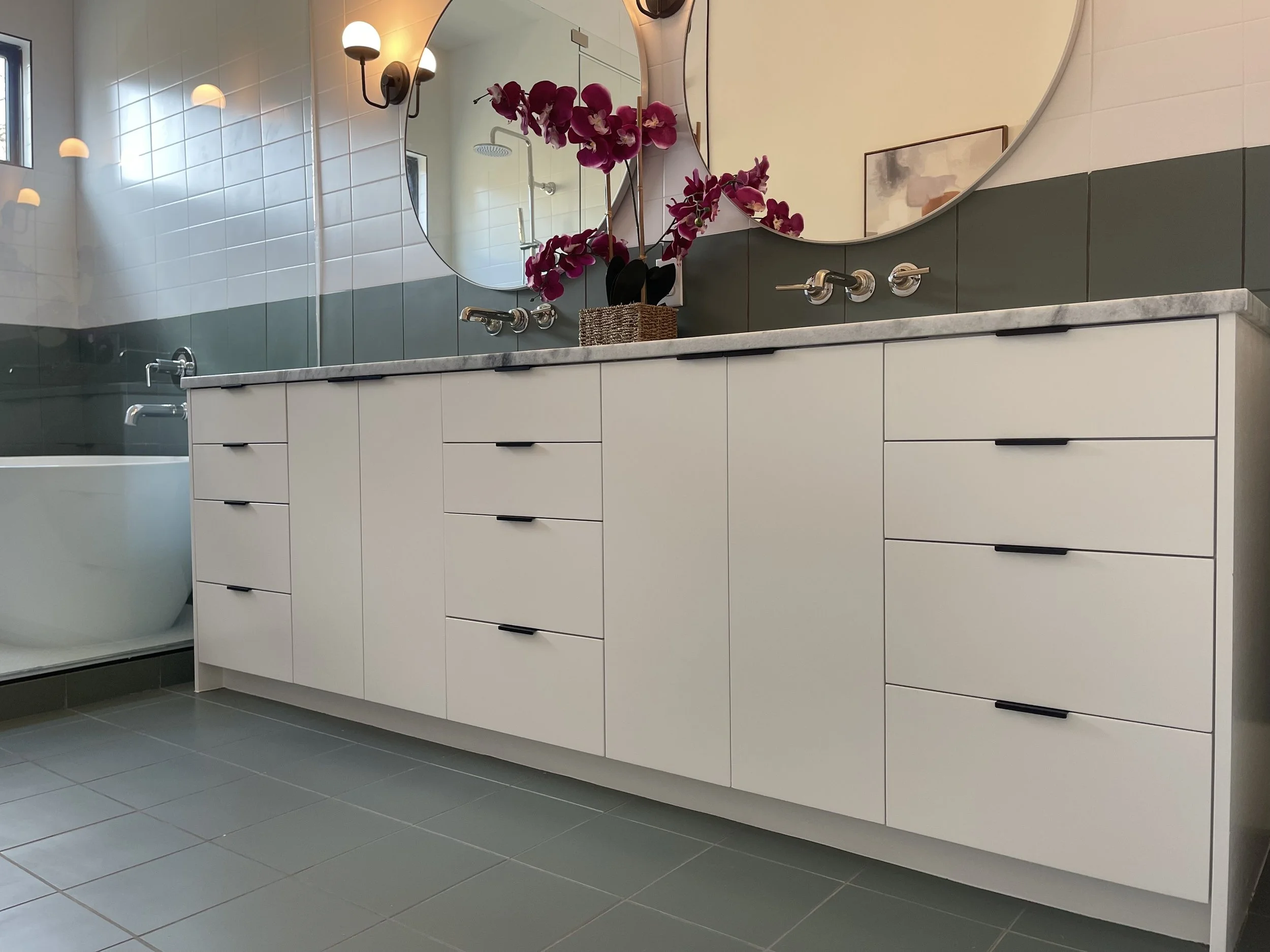 Modern bathroom with white cabinet vanity, marble countertop, large round mirror, purple orchids in a basket, wall-mounted faucets, and a bathtub with a glass shower door.