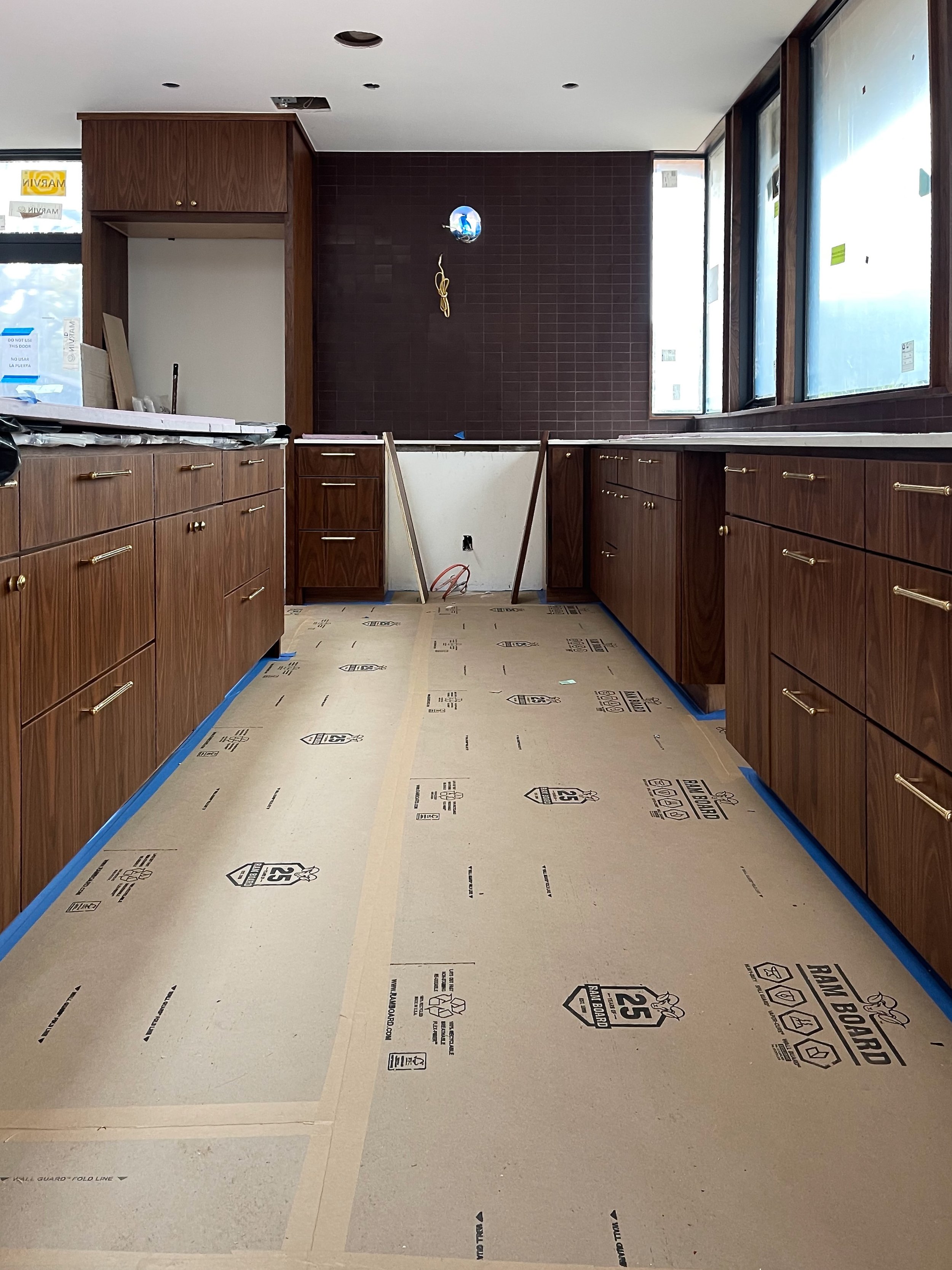 Kitchen under construction with brown wooden cabinets, protective floor covering, and large windows.