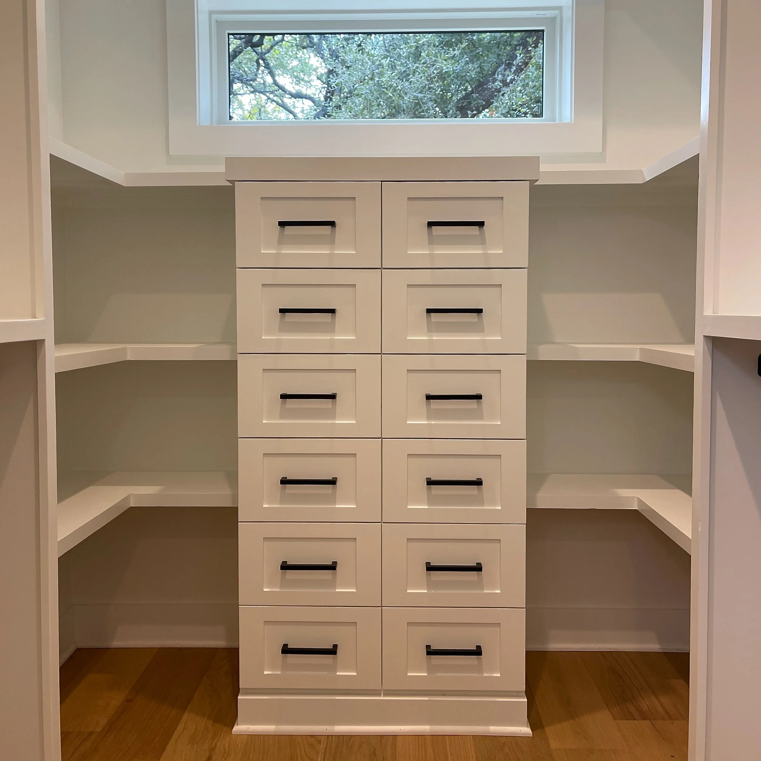 White built-in shelving unit with multiple drawers and open shelves, located beneath a window with trees outside.