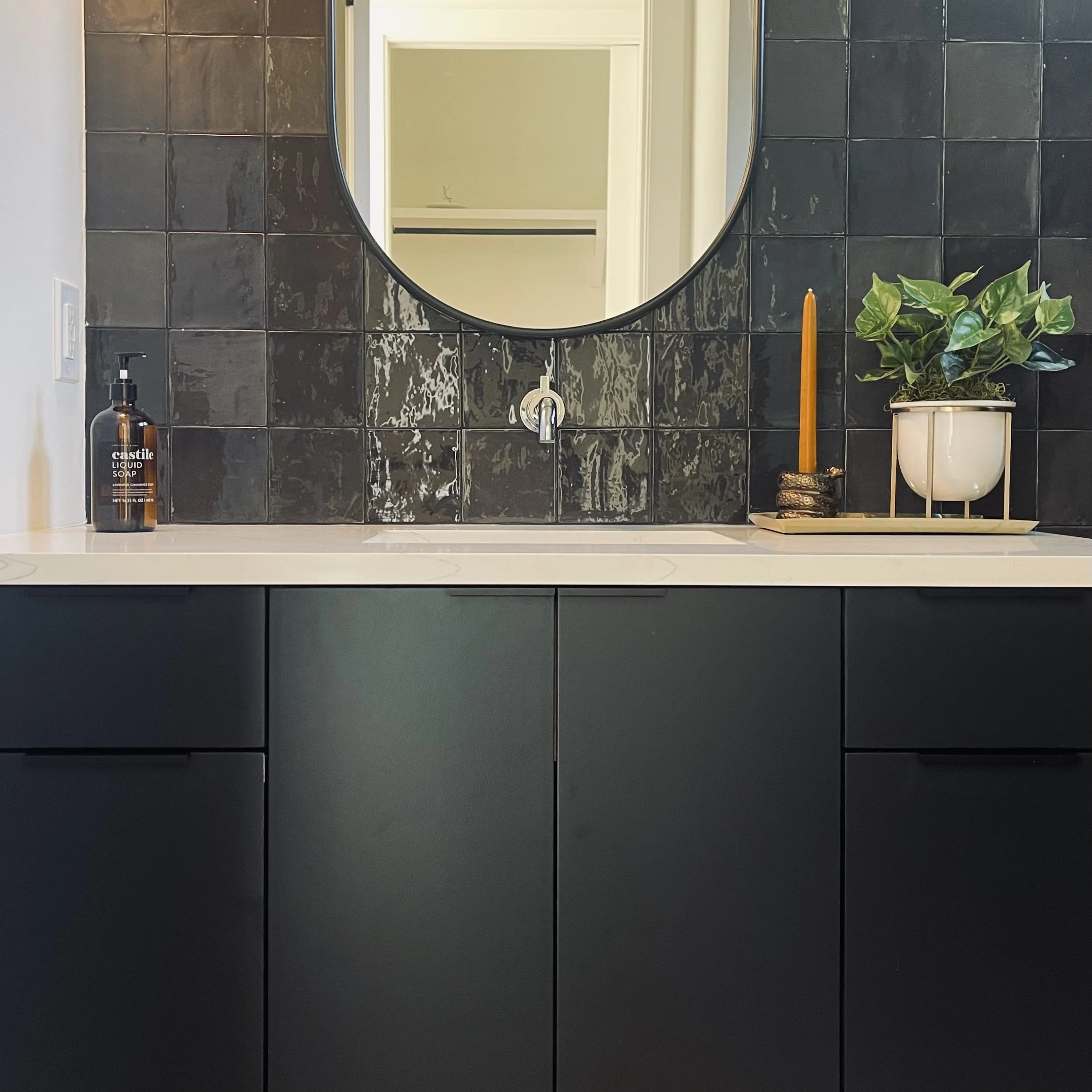 Bathroom vanity with black cabinet, white countertop, round mirror, black tiled wall, a bottle of liquid soap, a plant in a white pot, a candle, and a decorative tray.