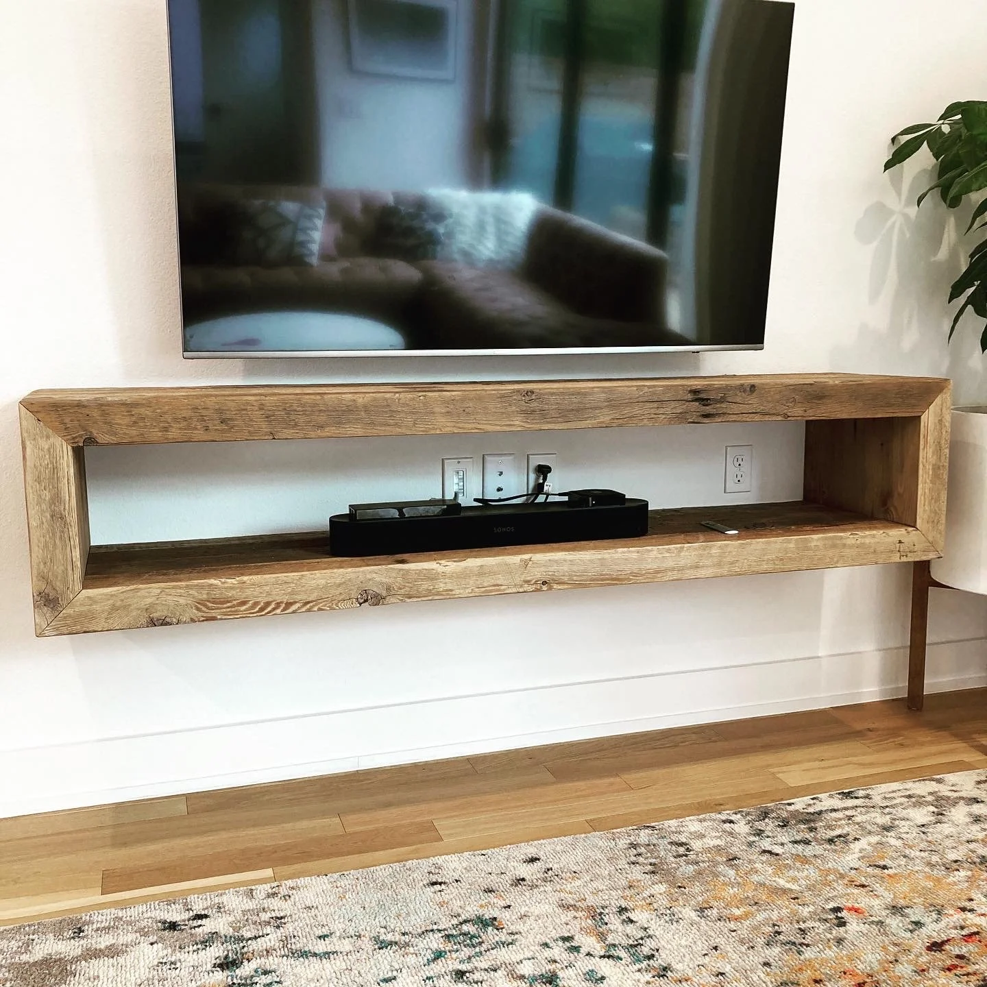 Wall-mounted flat-screen TV above a wooden shelf with a sound bar and electrical outlets, with a beige and brown patterned rug and a potted plant nearby.