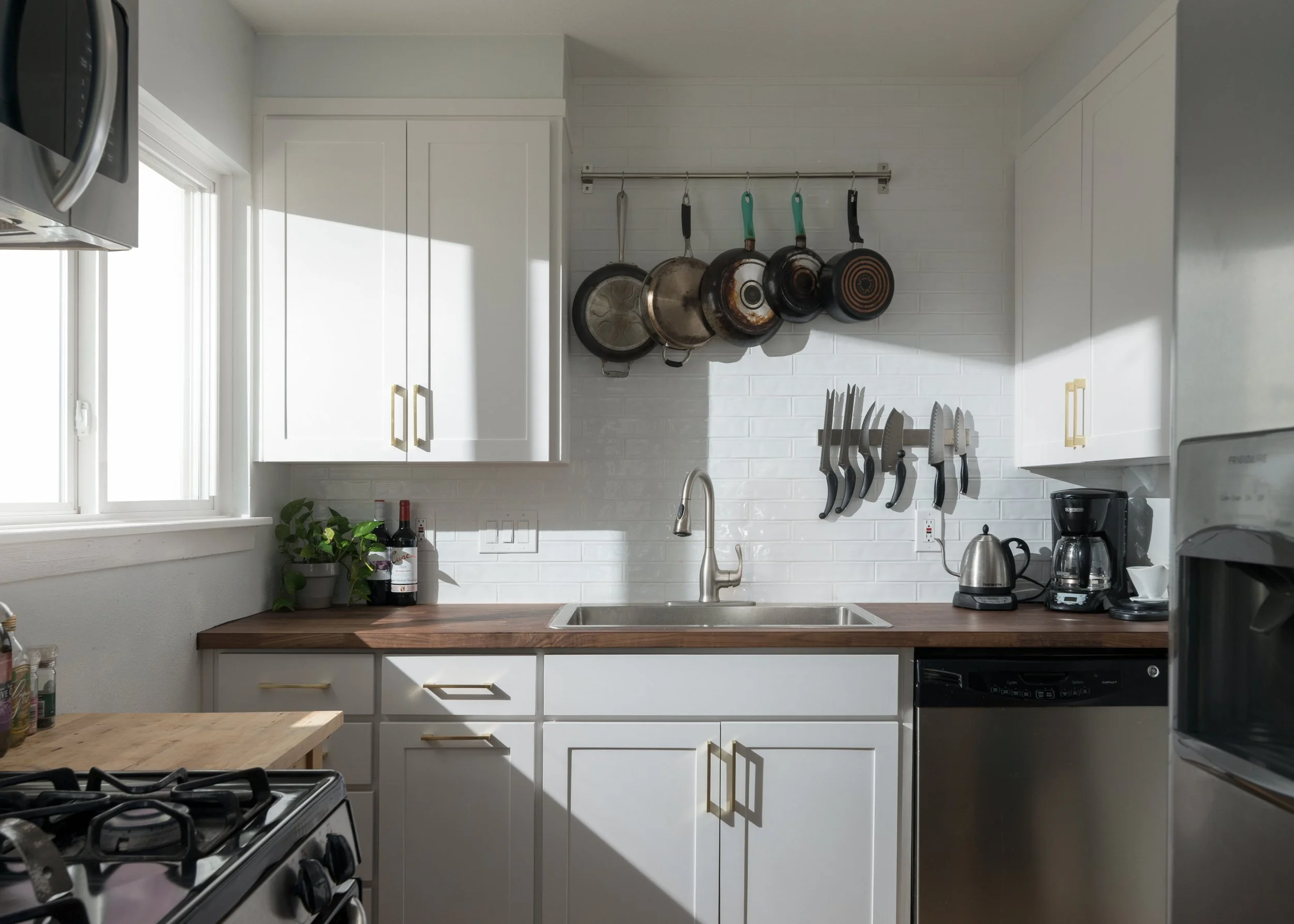 Modern kitchen with white cabinets, wooden countertop, stainless steel appliances, and hanging pots and knives, illuminated by natural sunlight.