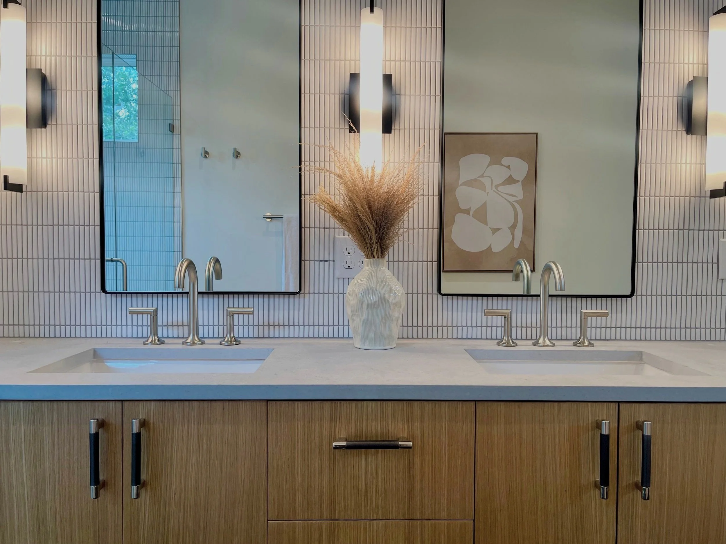 Bathroom with two sinks, two mirrors, a vase with dried plants, and a framed abstract art piece on beige tiled wall.