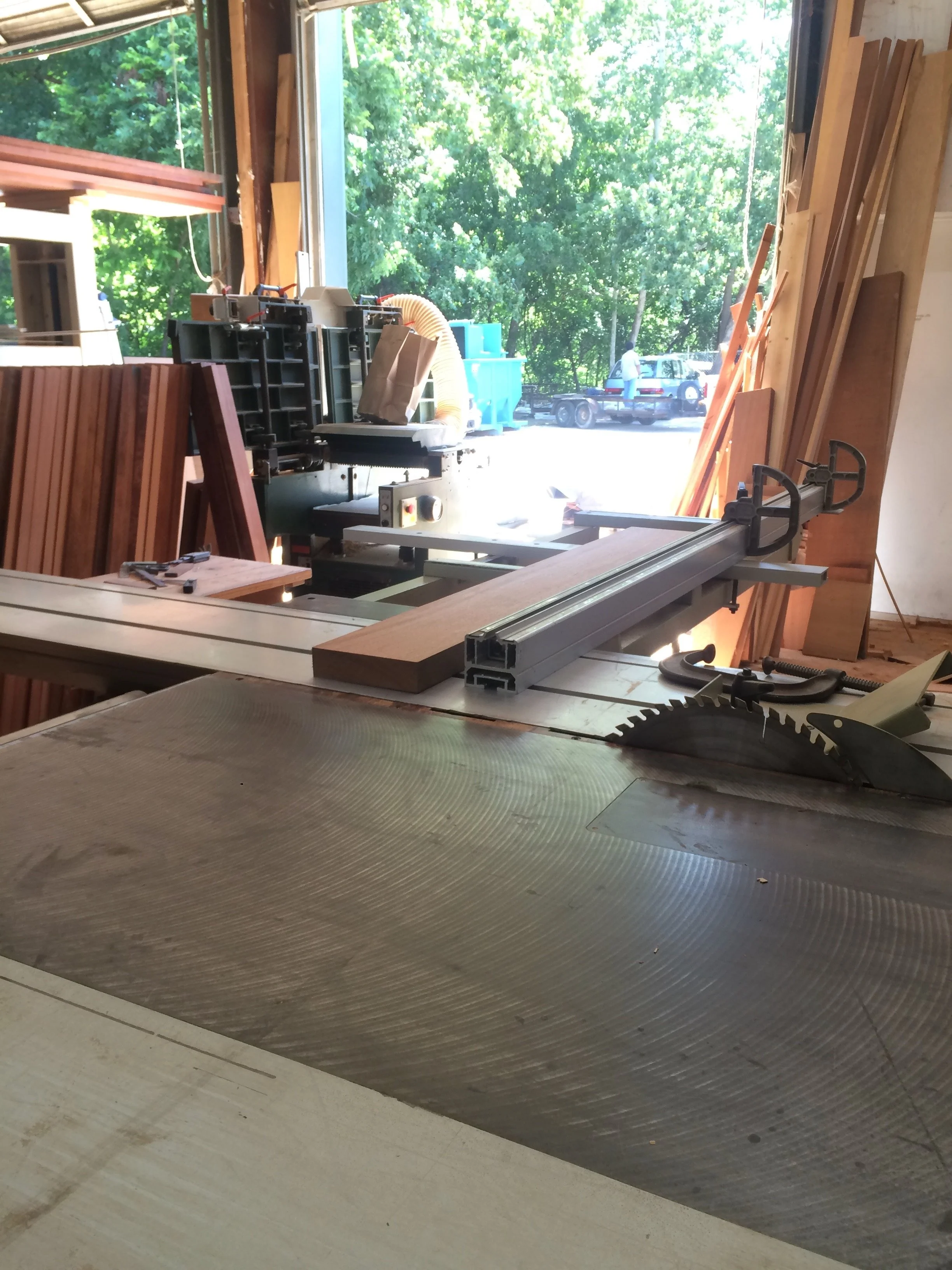 Interior view of a woodworking shop with a table saw, a miter saw, and stacks of wood, with a large open window showing green trees and parked cars outside.