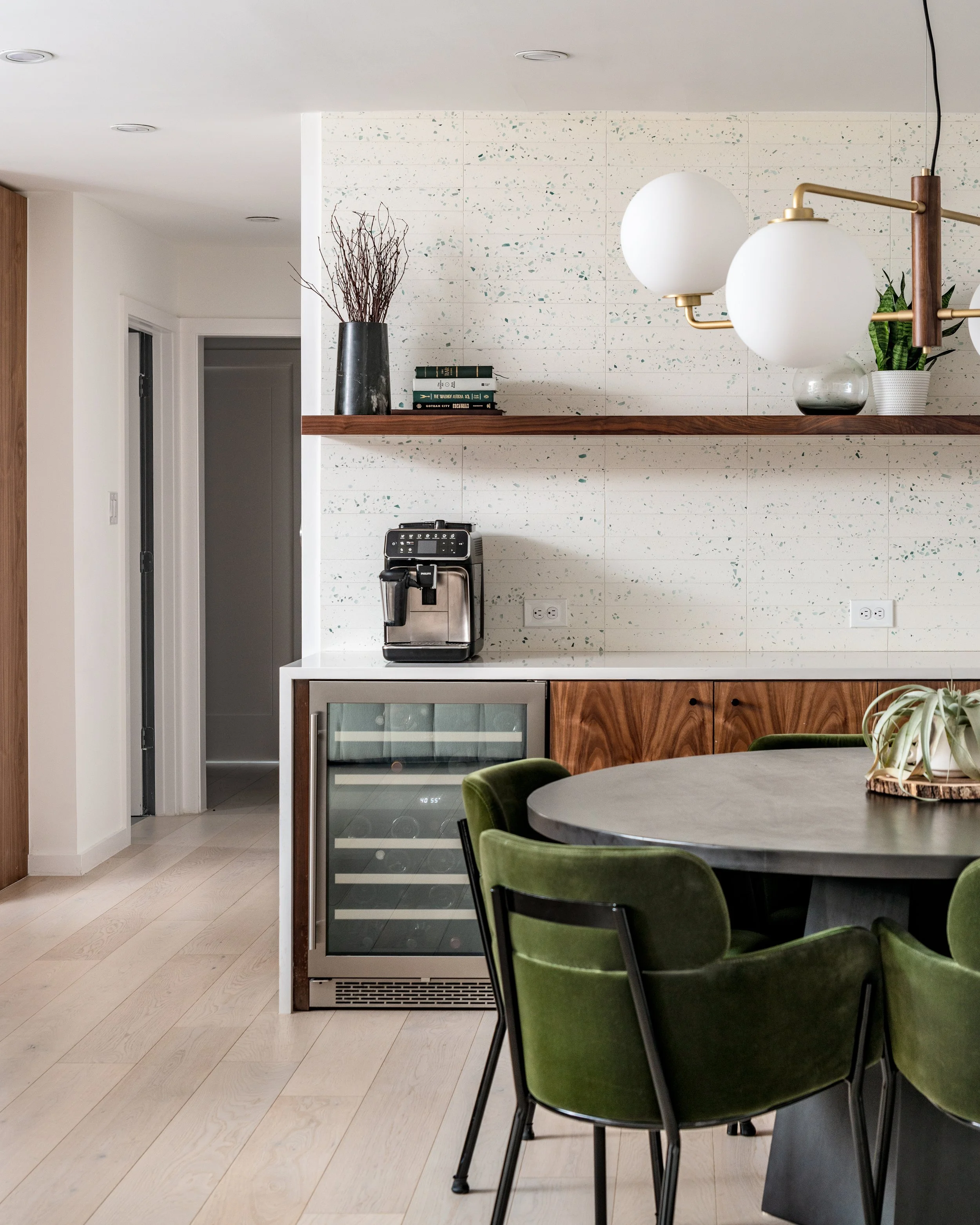 Interior view of a modern kitchen and dining area featuring a round table with green chairs, a built-in mini fridge, a coffee machine, and a wall-mounted shelf with decorative items and books.