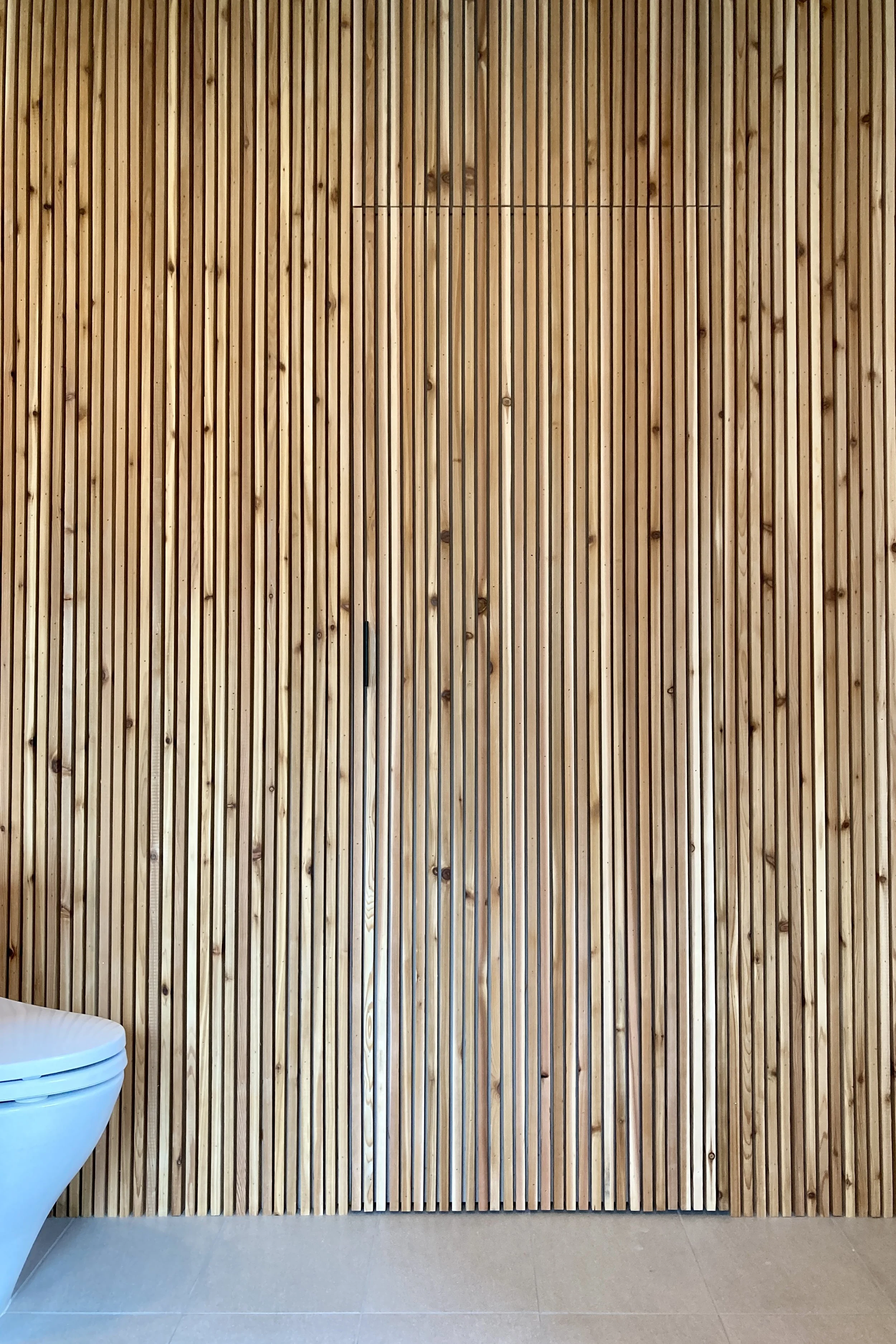 Wood-paneled wall with vertical slats in a modern interior, partial view of a white toilet on the left, light-colored tiled floor.