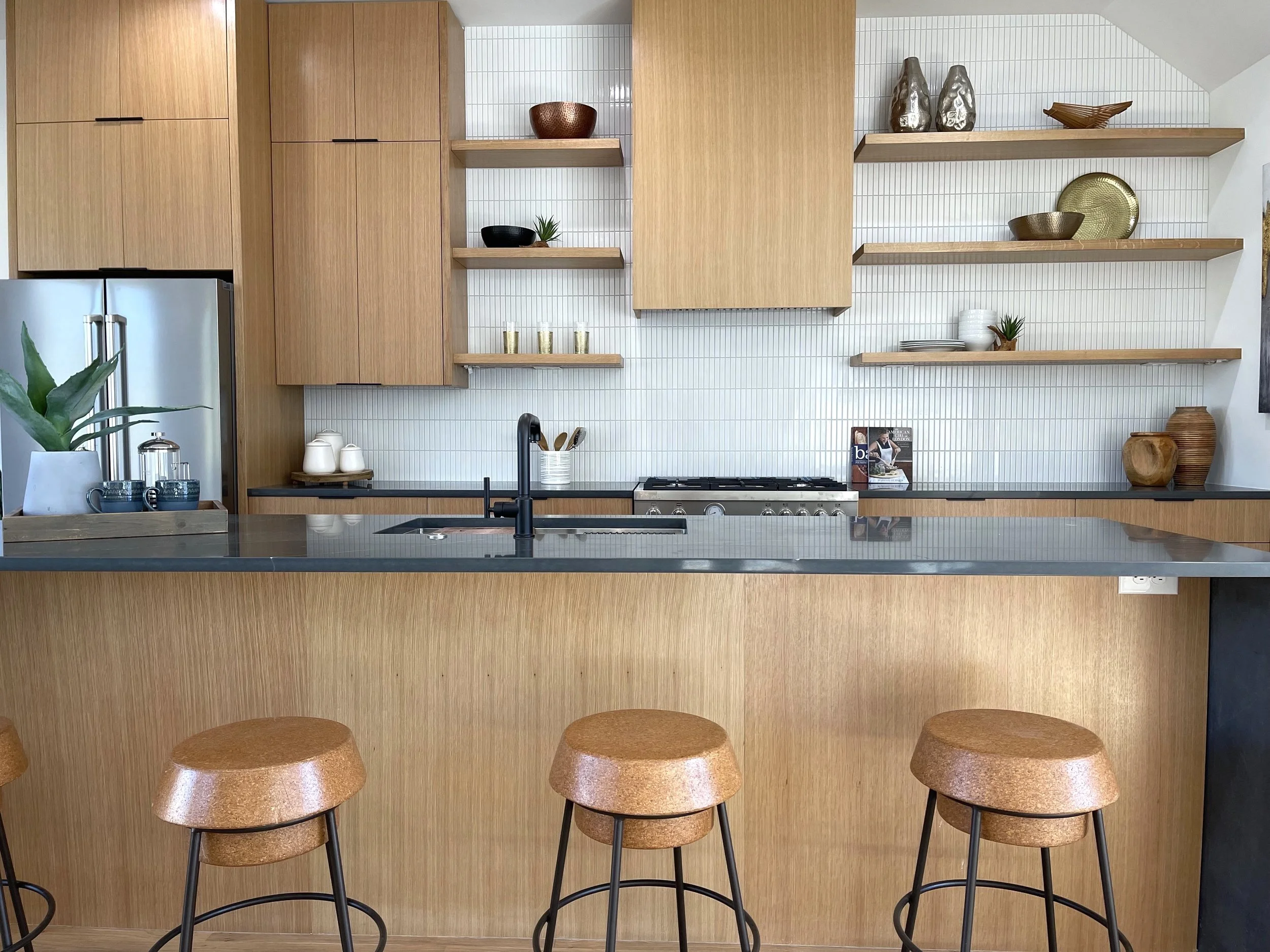 Modern kitchen with wooden cabinets, open shelves displaying decorative bowls and plates, a black faucet over a dark countertop, a small potted plant, and three brown stools at the island.