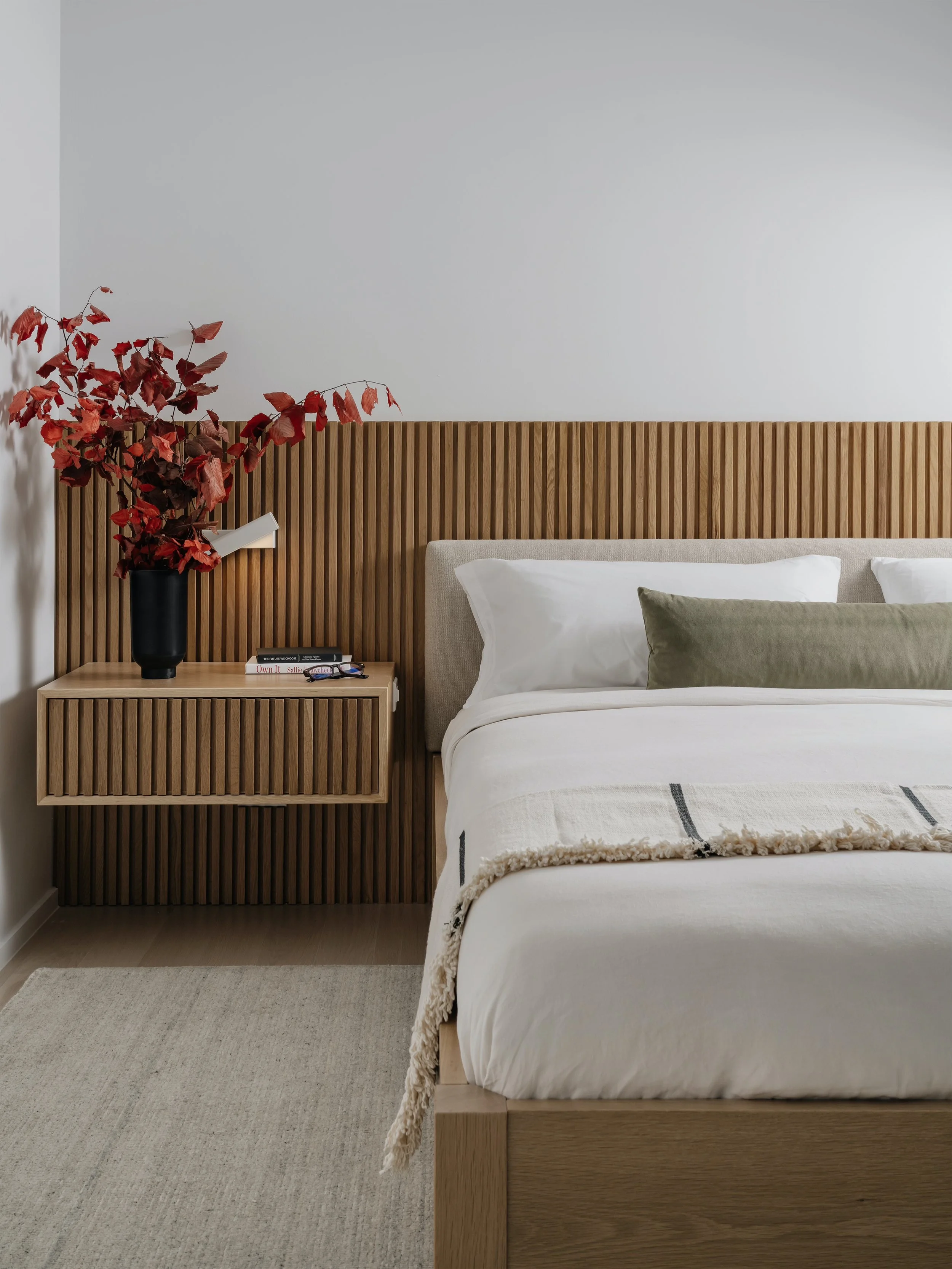 Modern bedroom with a wooden headboard, white bedding, green pillow, nightstand with black plant vase, red leaves, books, and glasses.