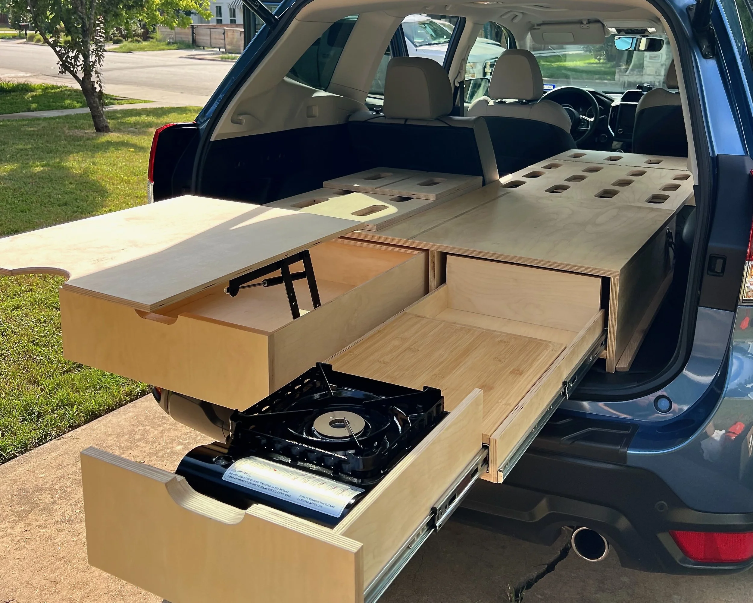 The back of a vehicle with custom wooden cabinetry and storage drawers, likely for camping or travel, with a built-in stove and counter space.