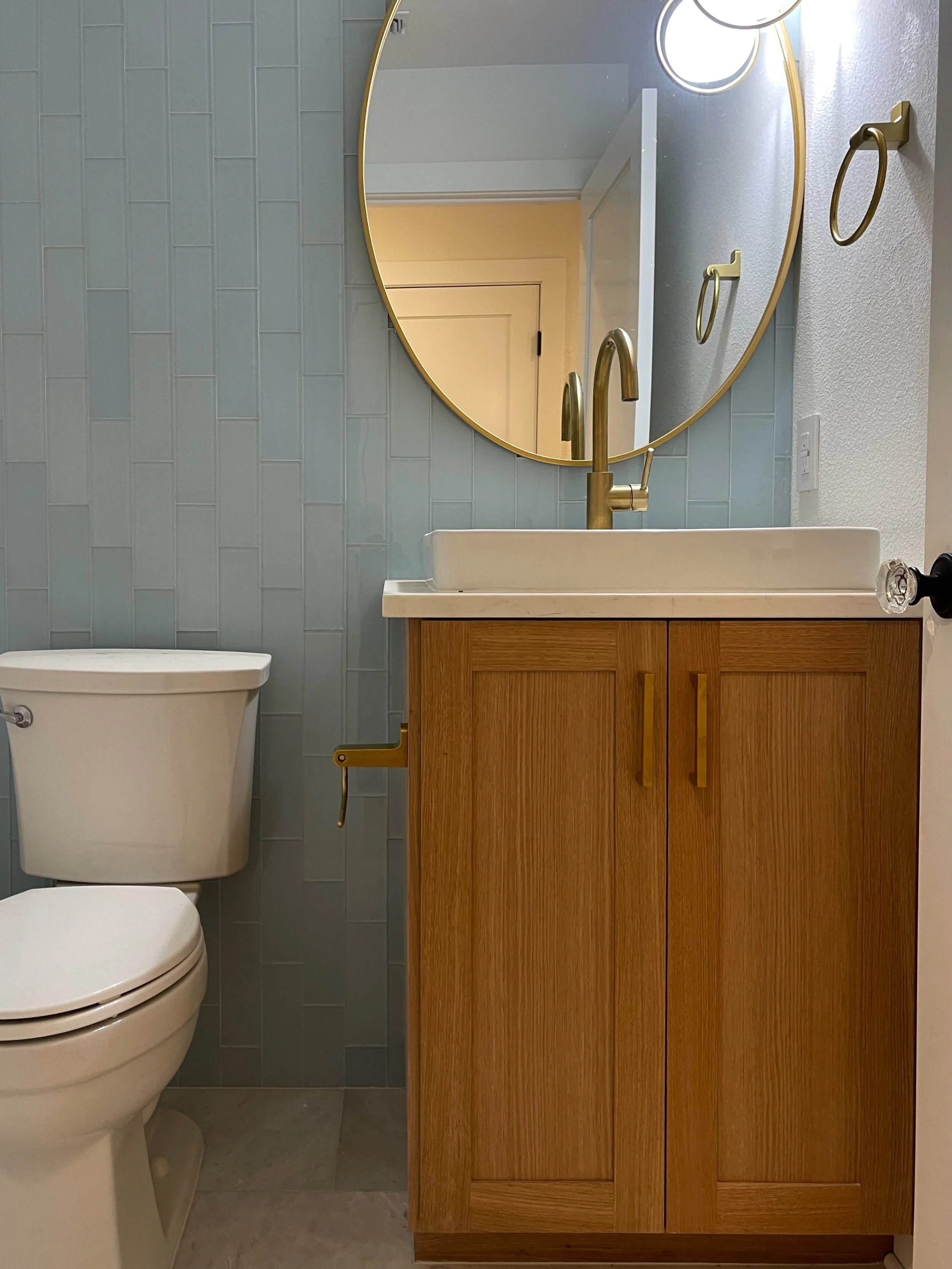 Bathroom with a toilet, a wooden vanity with a white sink, and an oval mirror on a textured gray tile wall.