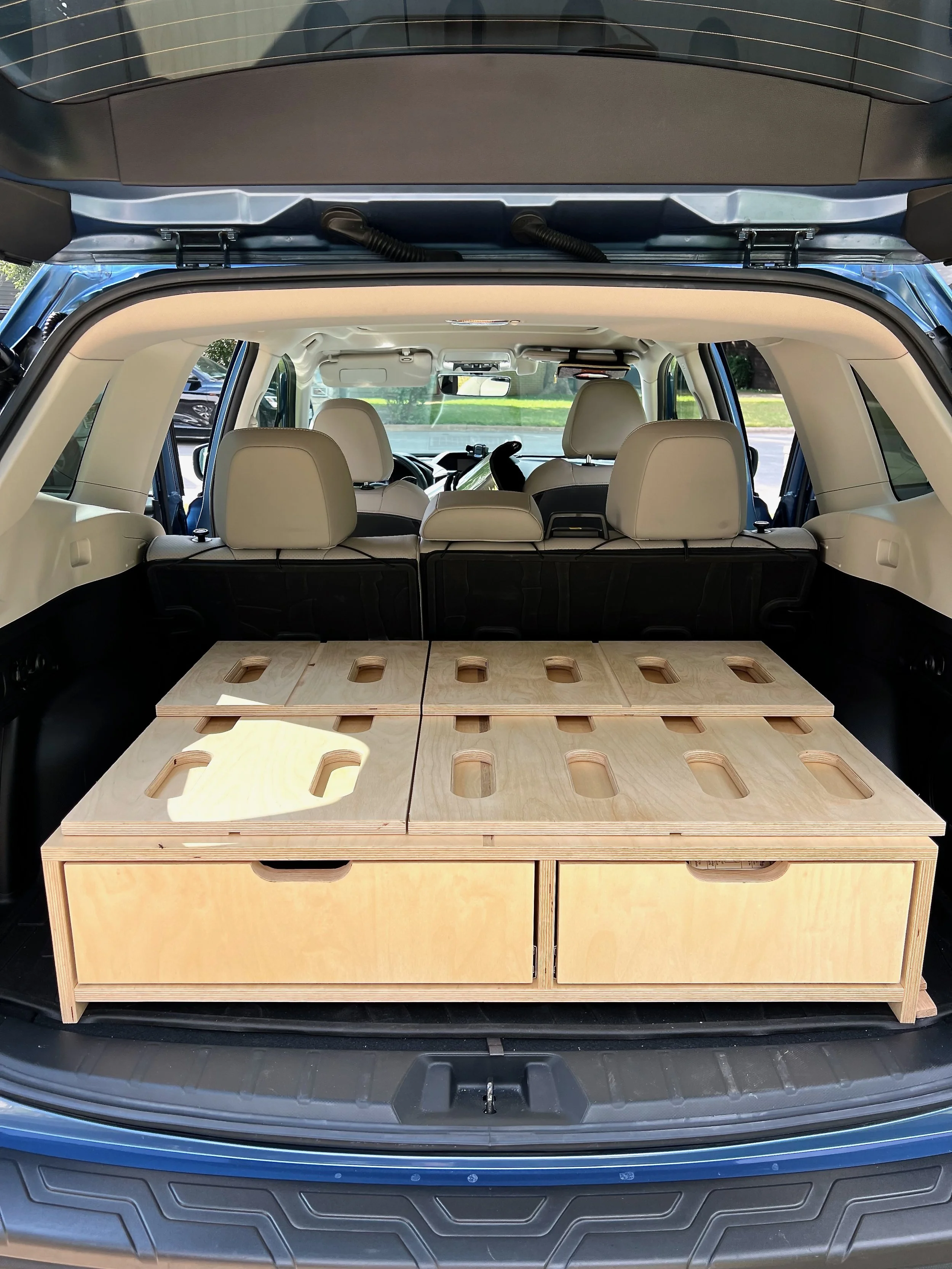 The trunk of an SUV with a custom wooden storage unit installed, featuring multiple compartments and cutouts, with beige seats and a view of the front interior visible.