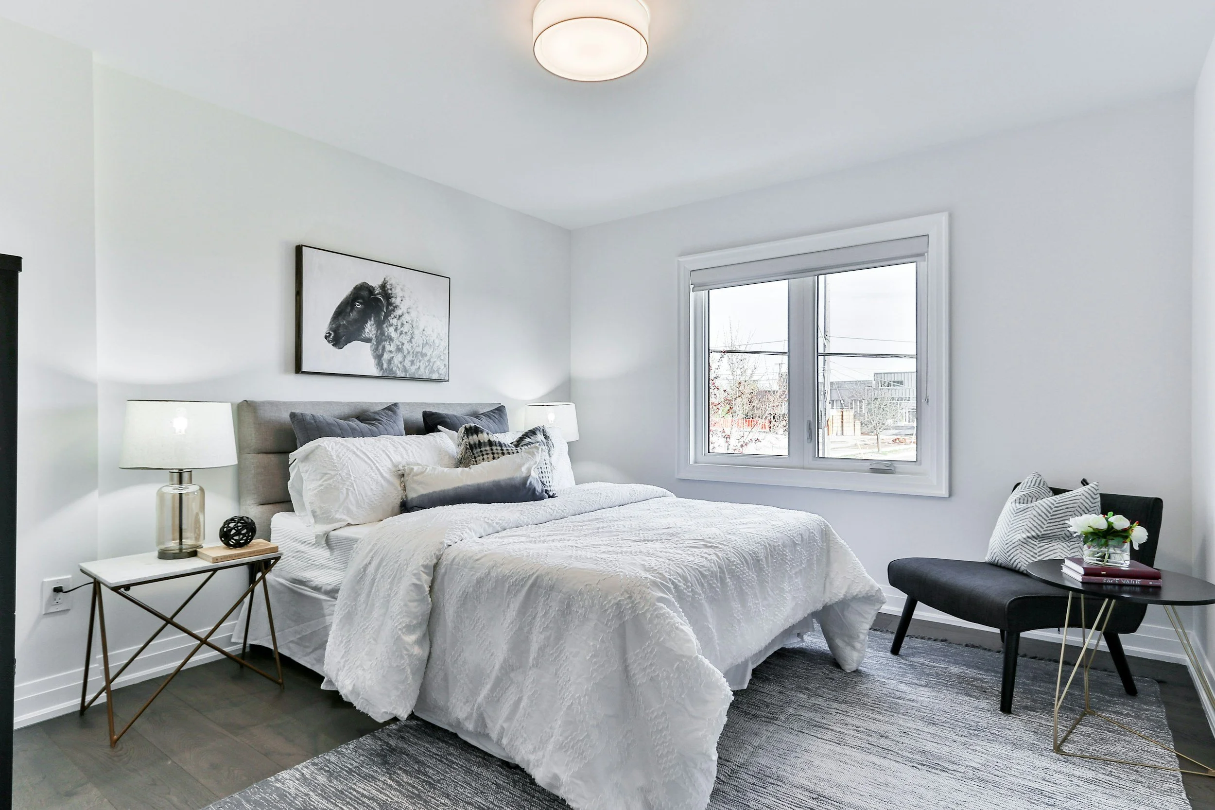 A modern bedroom with white walls, a bed with white bedding and gray pillows, a black and white sheep artwork above the bed, a nightstand with a glass lamp, a window with a view of a snowy neighborhood, and a black chair with a bow pillow and a flower vase on a side table.