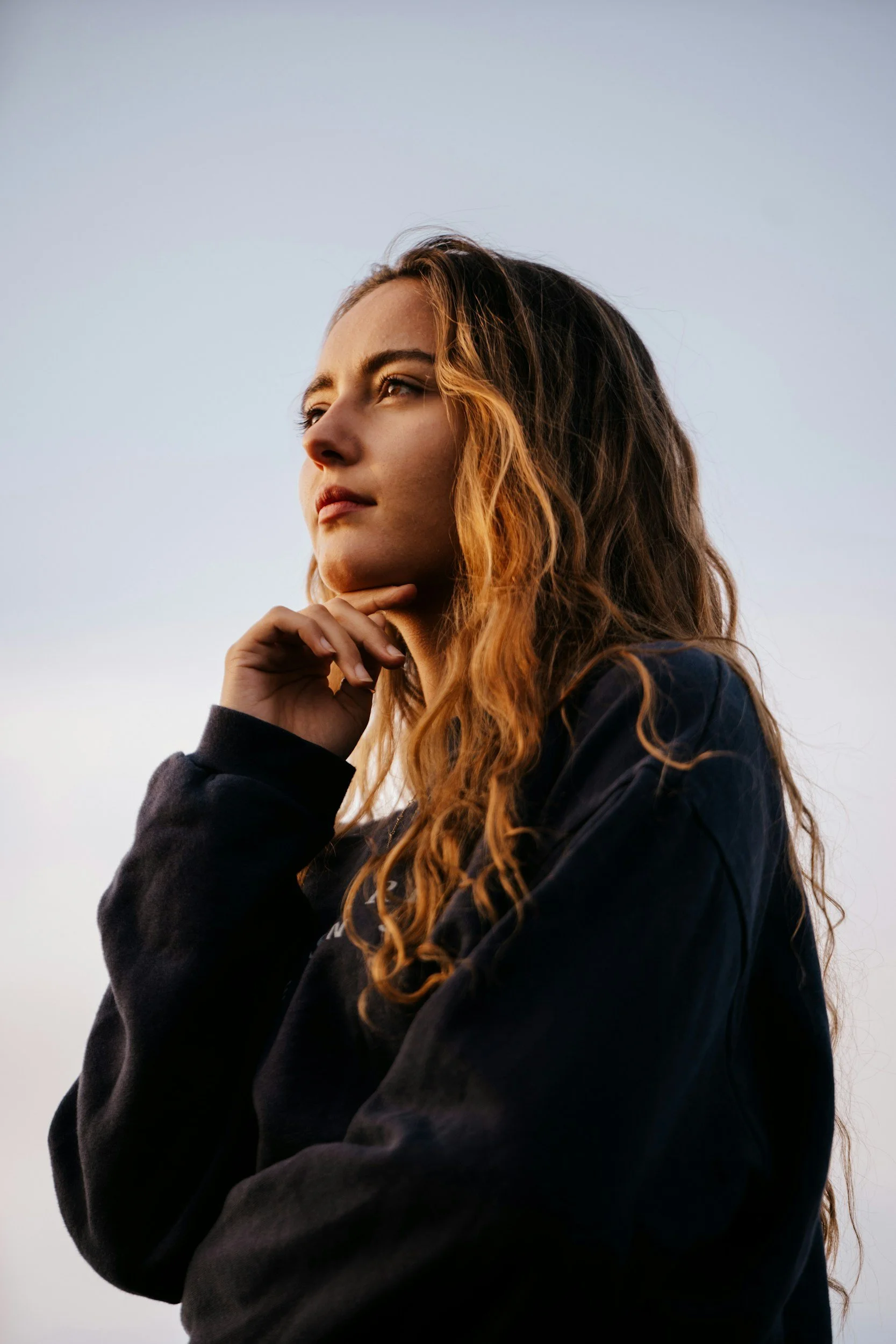A young woman with light skin and long, curly auburn hair standing outdoors against a pale sky, touching her chin with her right hand, wearing a black hoodie.