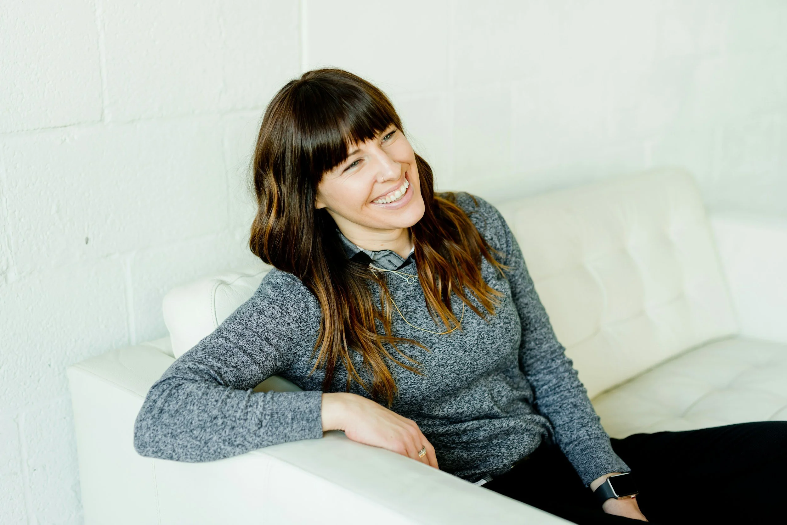 A woman with long brown hair and bangs, smiling, sitting on a white couch against a white brick wall, wearing a gray patterned sweater and a smartwatch.