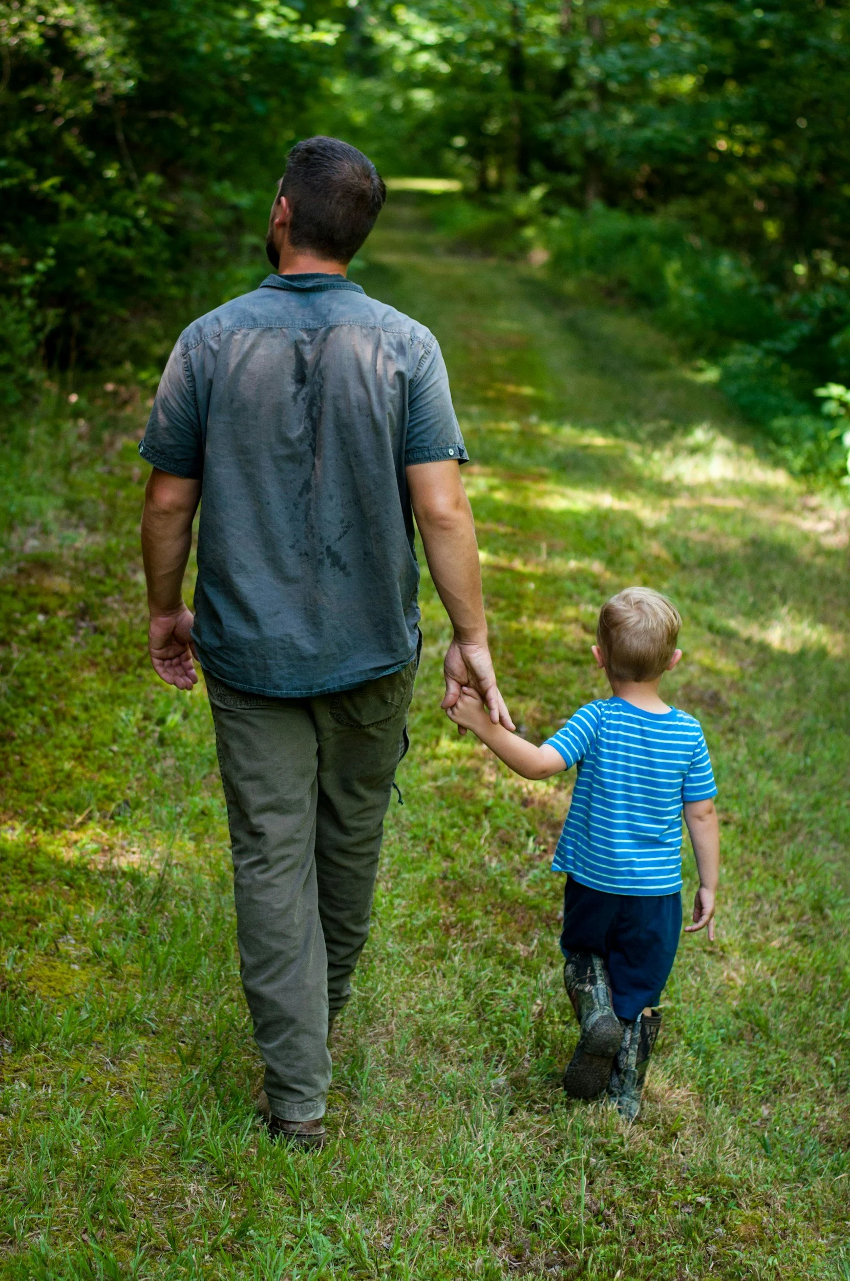 An adult man and a young boy walk hand-in-hand down a grassy forest trail surrounded by green foliage.