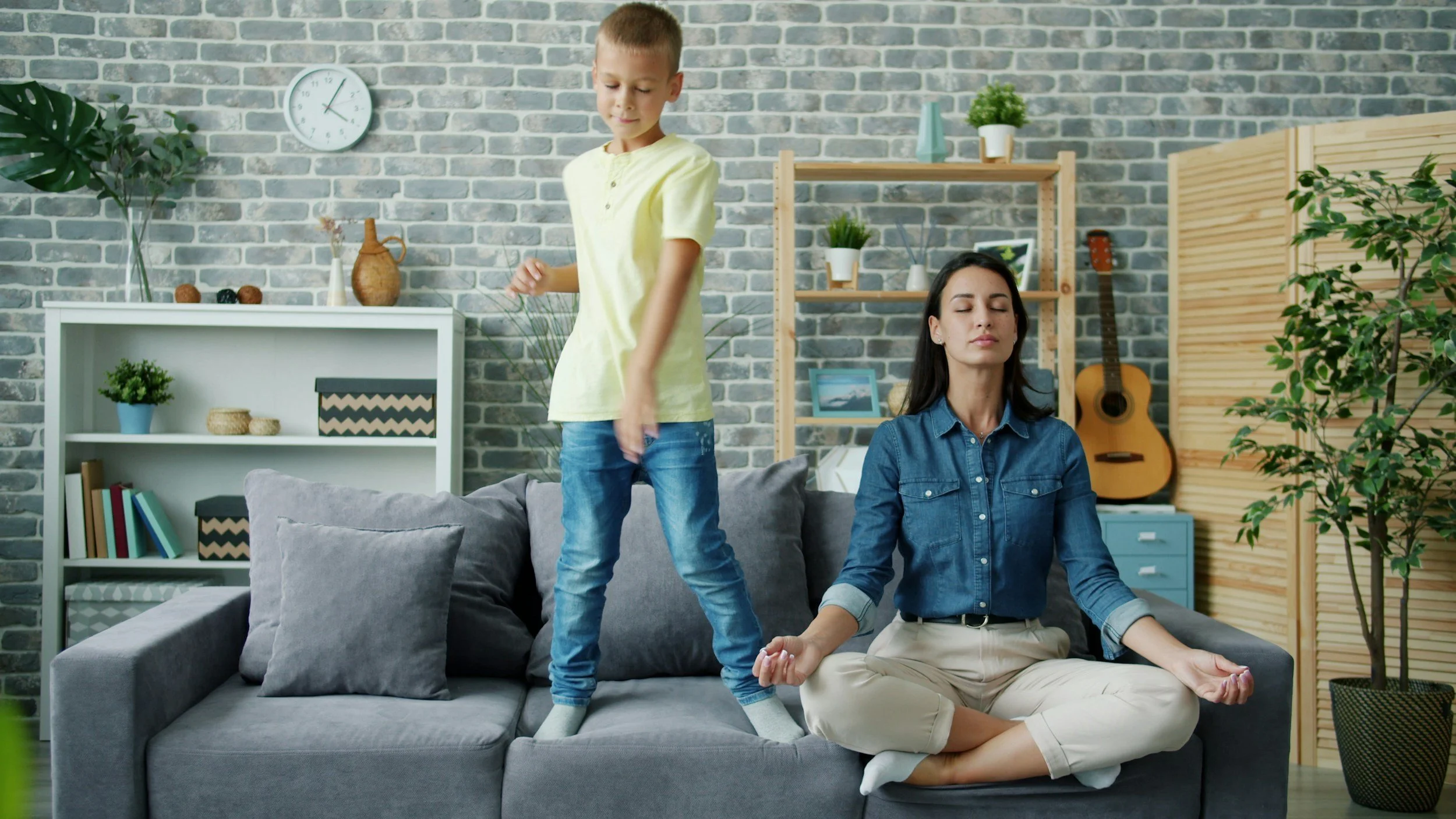 A boy standing on a sofa with eyes closed and a woman sitting cross-legged on the sofa in a meditation pose with eyes closed, in a cozy living room with green plants and decorative items.