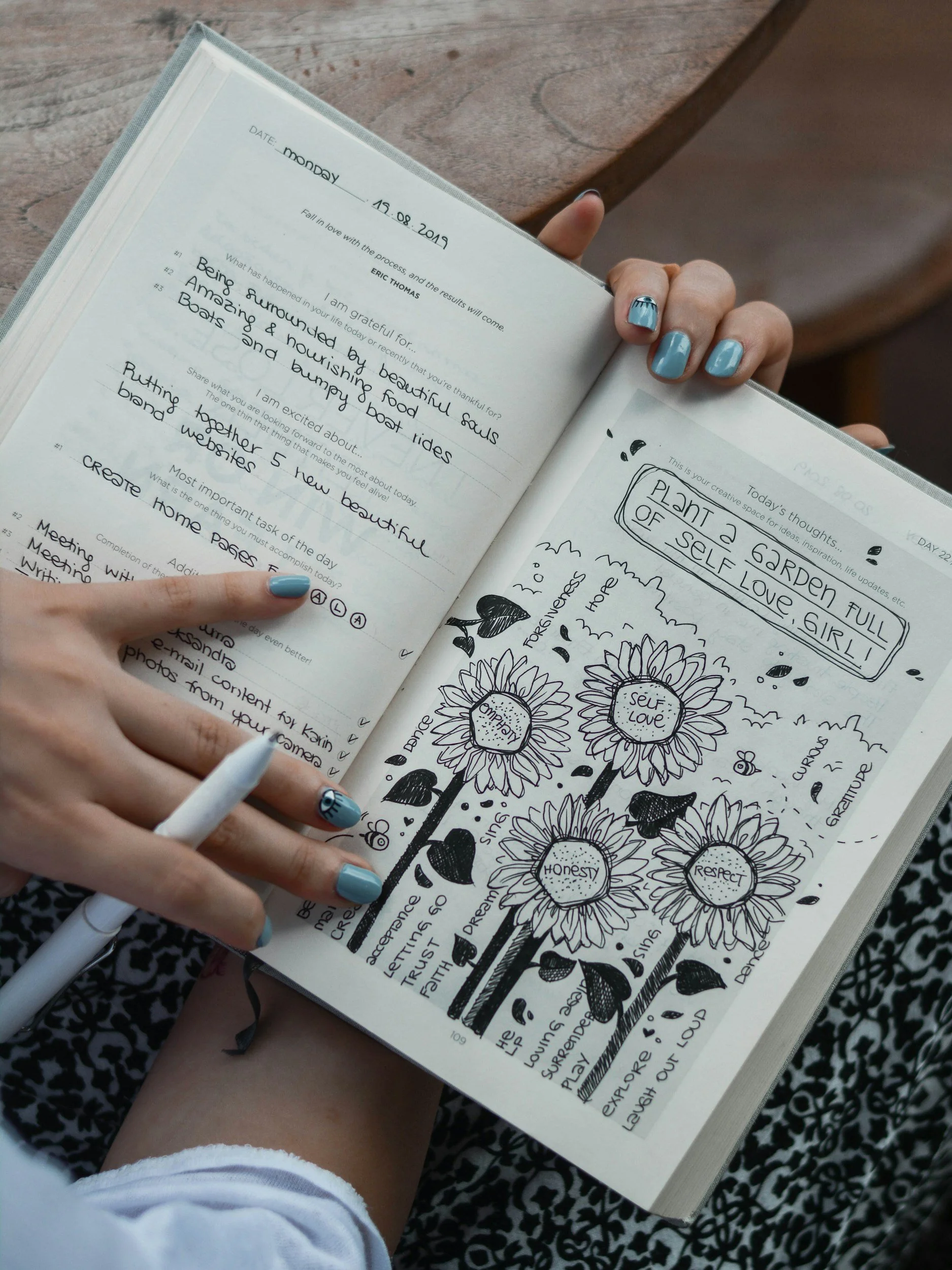 A person with painted nails holding an open black-and-white illustrated journal with handwritten notes and drawings, including sunflowers with words like 'Self Love,' 'Honesty,' and 'Respect.' The journal contains reflections, lists, and creative doodles.