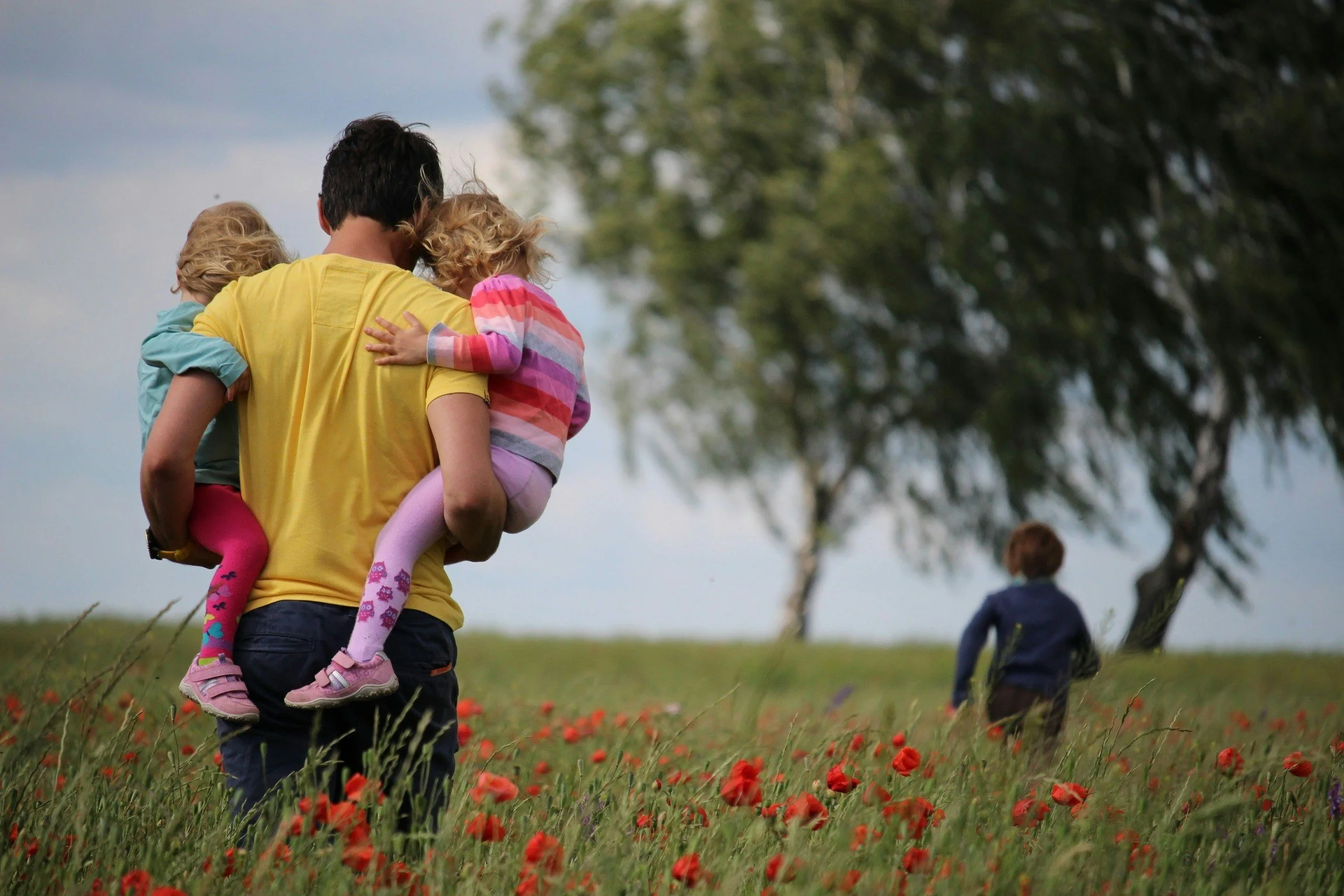 A man carrying two young children on his shoulders in a field of red poppies, with trees and a cloudy sky in the background.