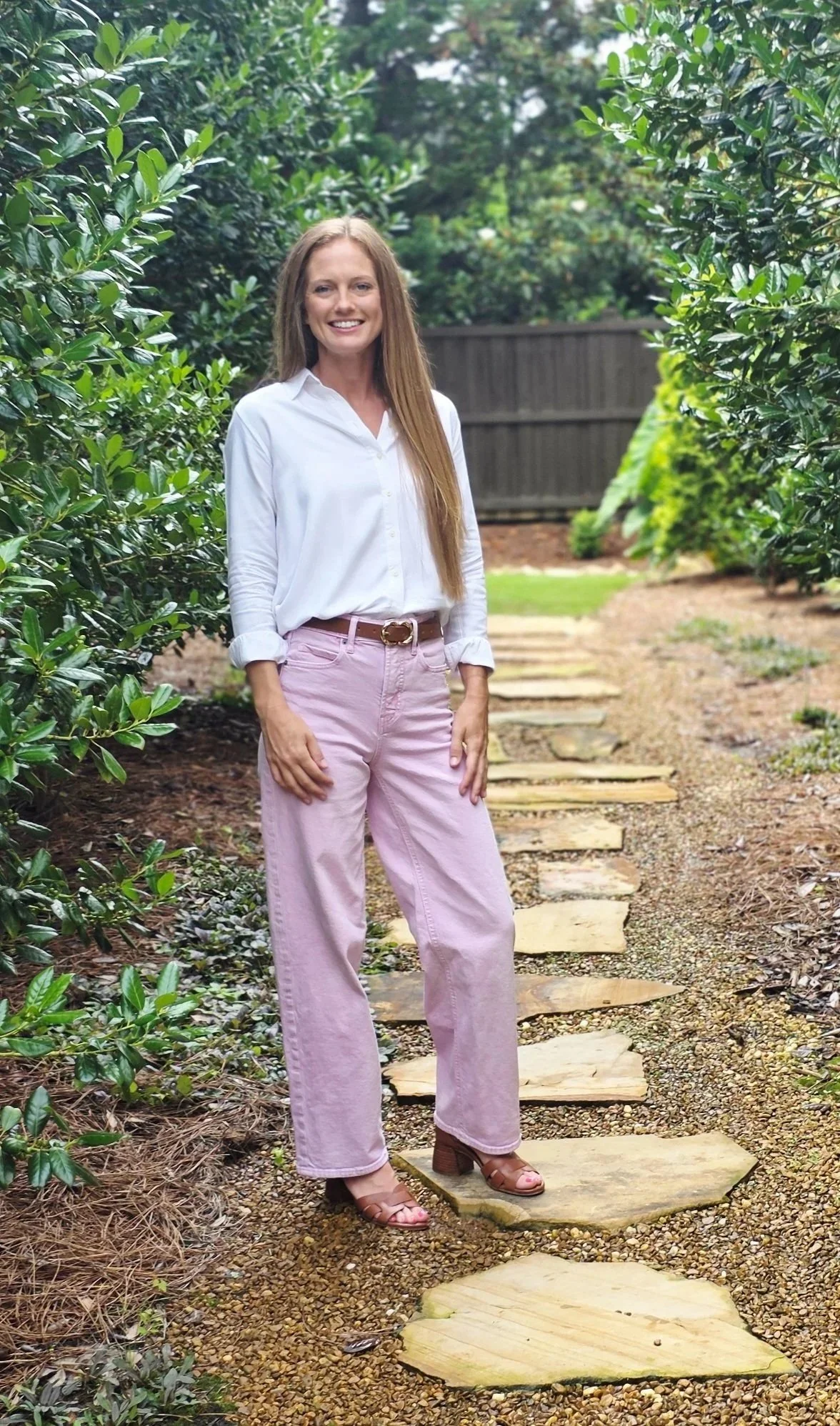 Woman with long hair smiling and standing on a stone pathway surrounded by green bushes and trees, wearing a white blouse, pink pants, and brown sandals.