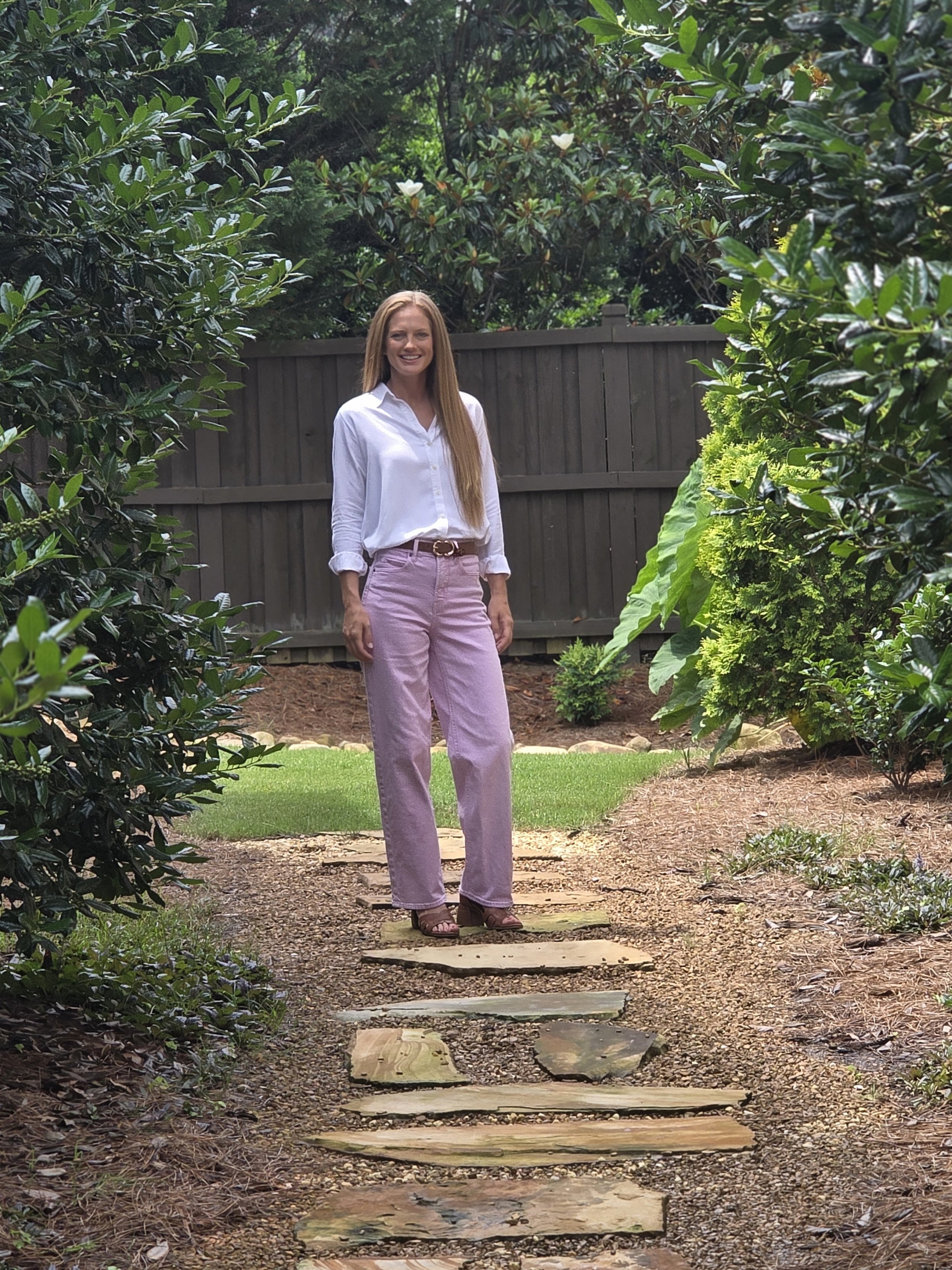 A woman smiling and standing on a stone pathway in a garden surrounded by green bushes and trees.