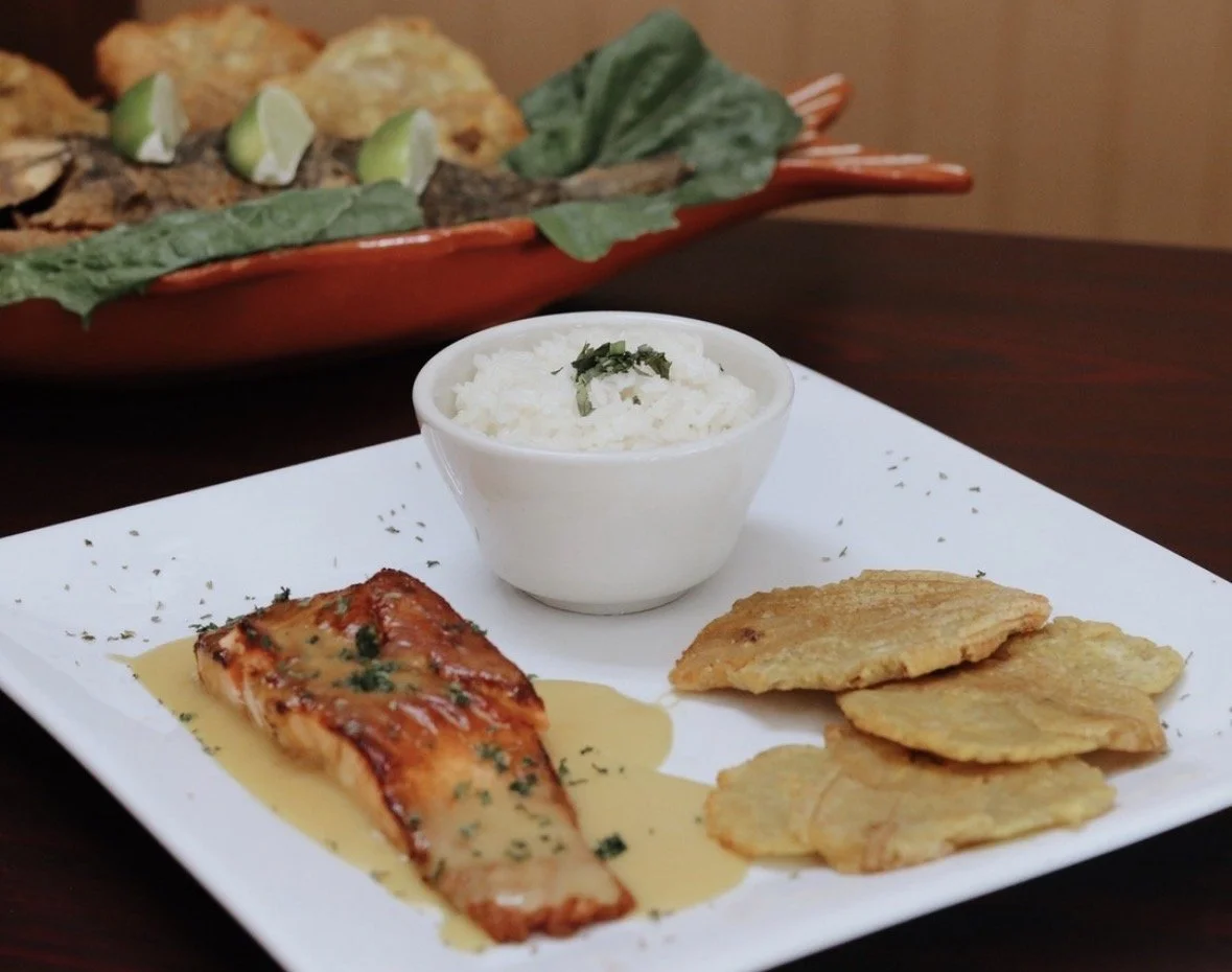 Plate of grilled fish, white rice with garnish, and fried plantain on a white square plate.