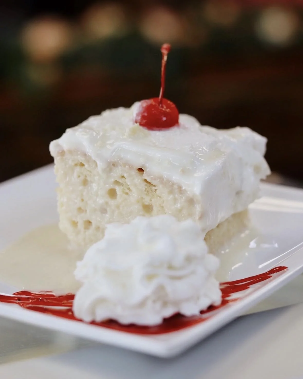A slice of tres leches cake with white frosting topped with a cherry, served with whipped cream and red syrup on a white plate.