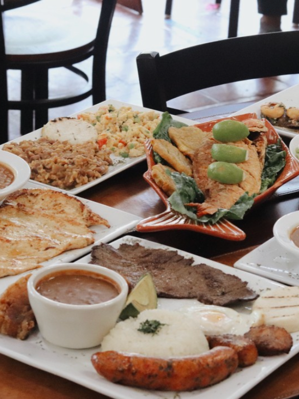 Assorted Colombian dishes on a table, including fried fish, rice, beans, grilled meat, a cooked egg, on a serving platter, with side dishes and sauces.