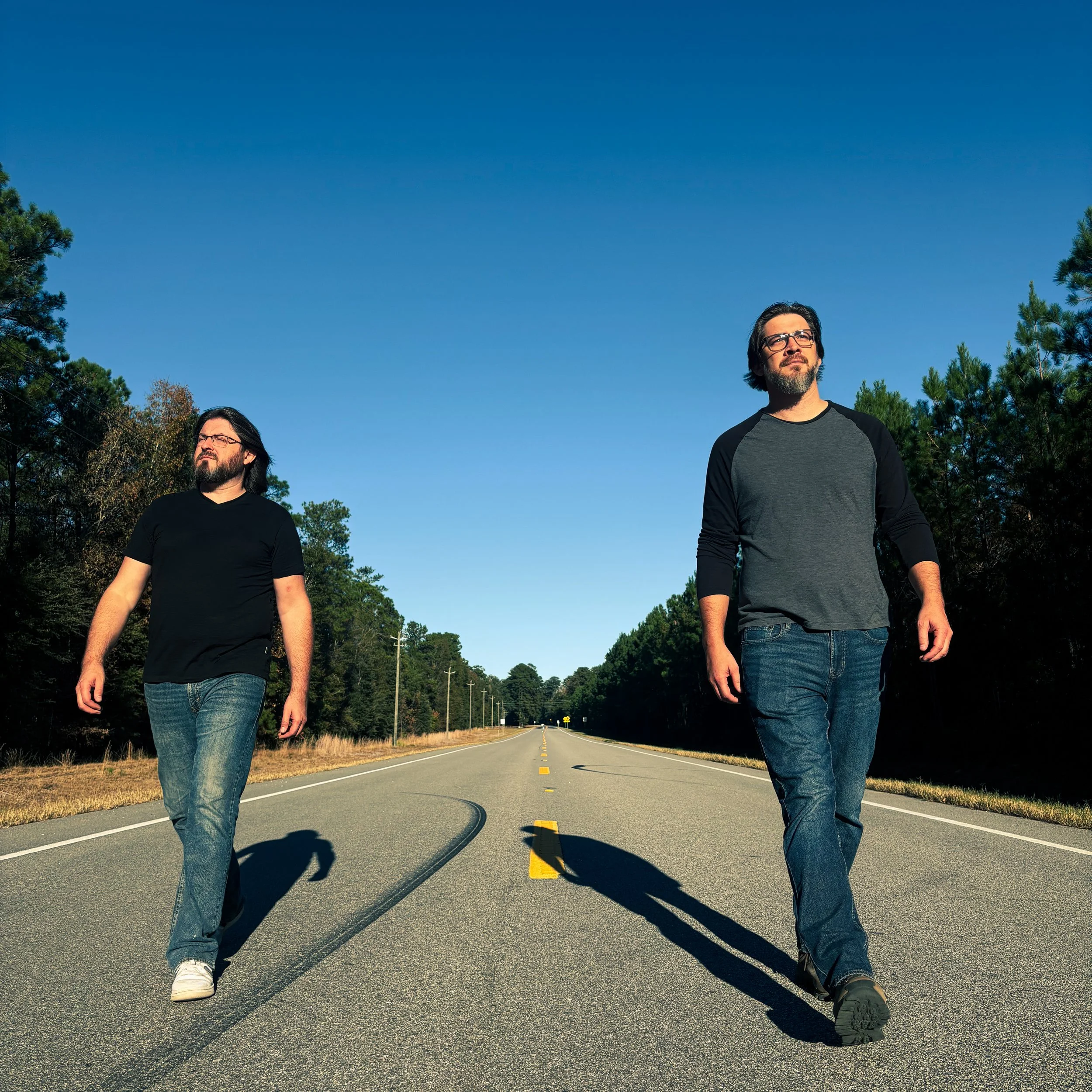 Two men walking on a rural road with a clear blue sky, surrounded by trees.