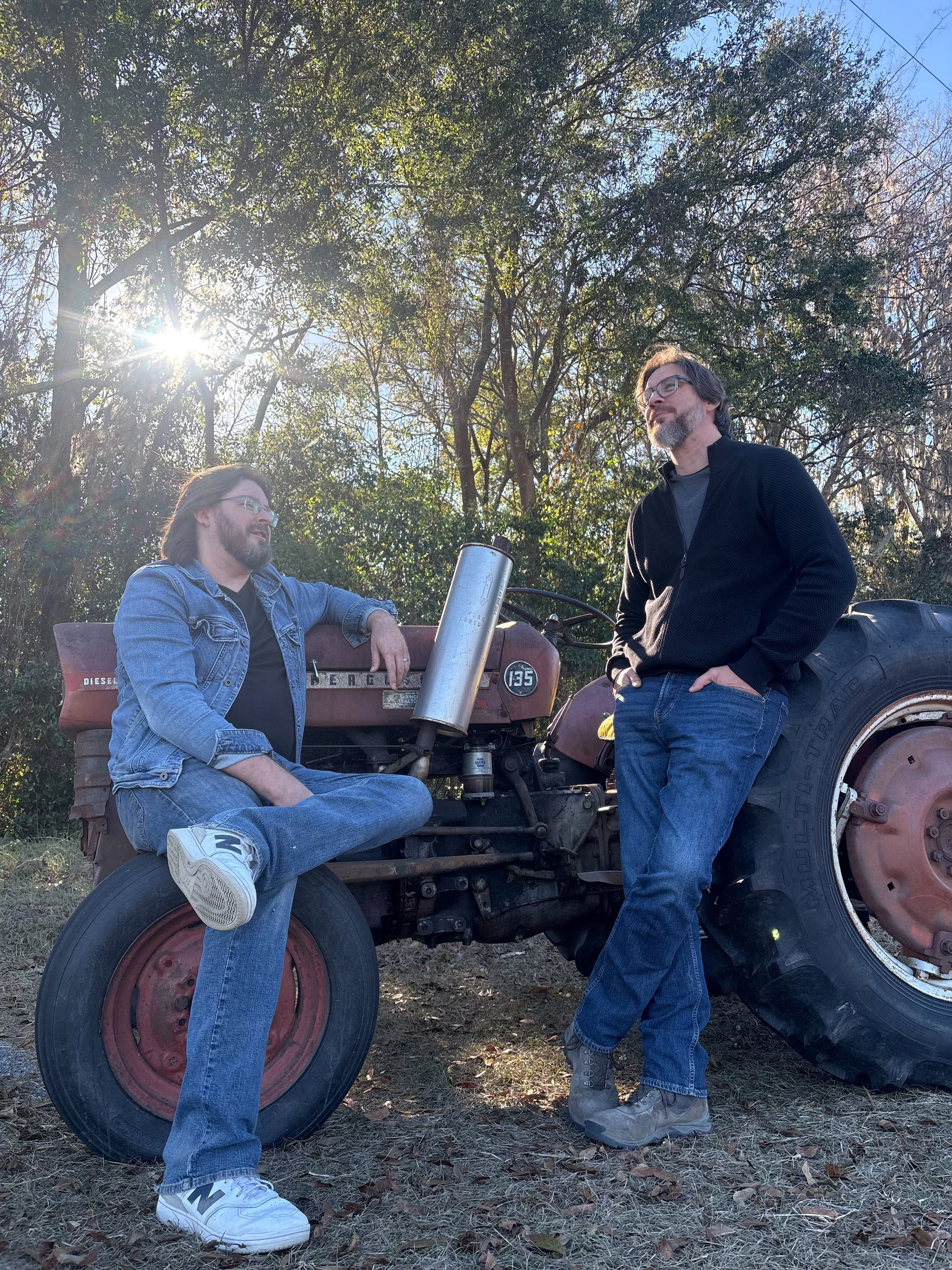Two men talking outdoors with an old tractor and trees in the background, sunlight shining through the trees, one sitting on the tractor, the other standing beside it.
