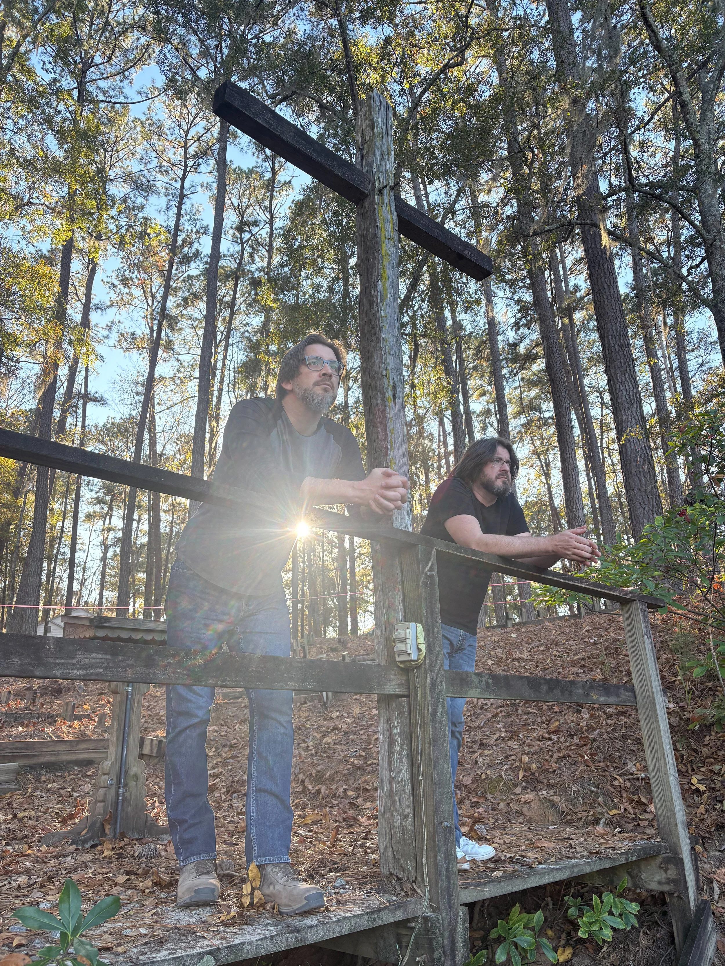 Two men standing on a wooden bridge outdoors in a forest, looking serious. One man has glasses, a beard, and curly hair, wearing a gray shirt. The other man has long hair and a beard, wearing a black shirt. The sun is shining behind them.