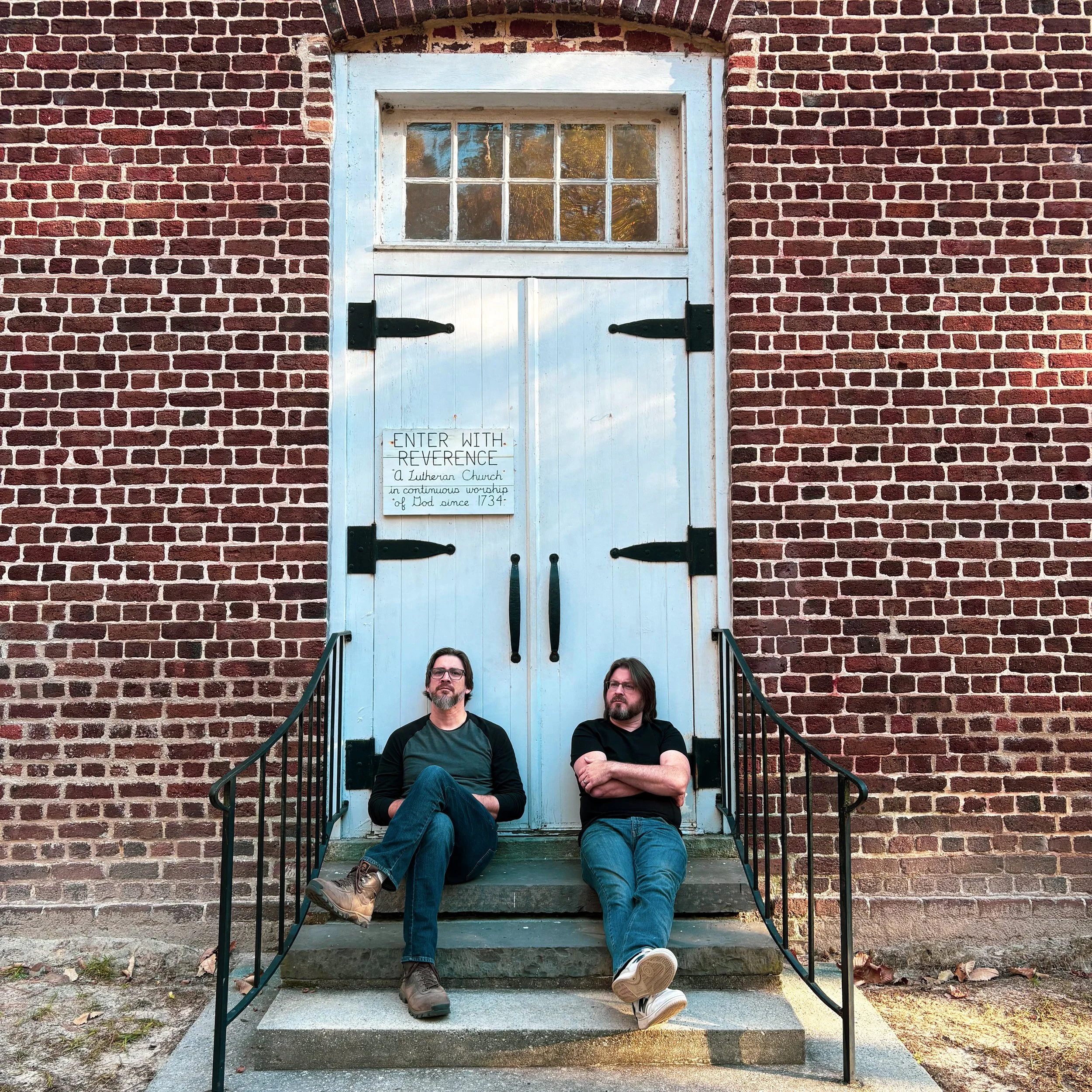 Two men sitting on the steps of a brick building with a white door and small window above, with a sign on the door that reads "Enter with reverence."