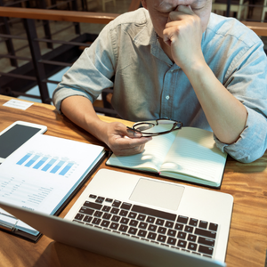 Person sitting at a table with a tablet, laptop, and notebook, holding glasses and thinking.