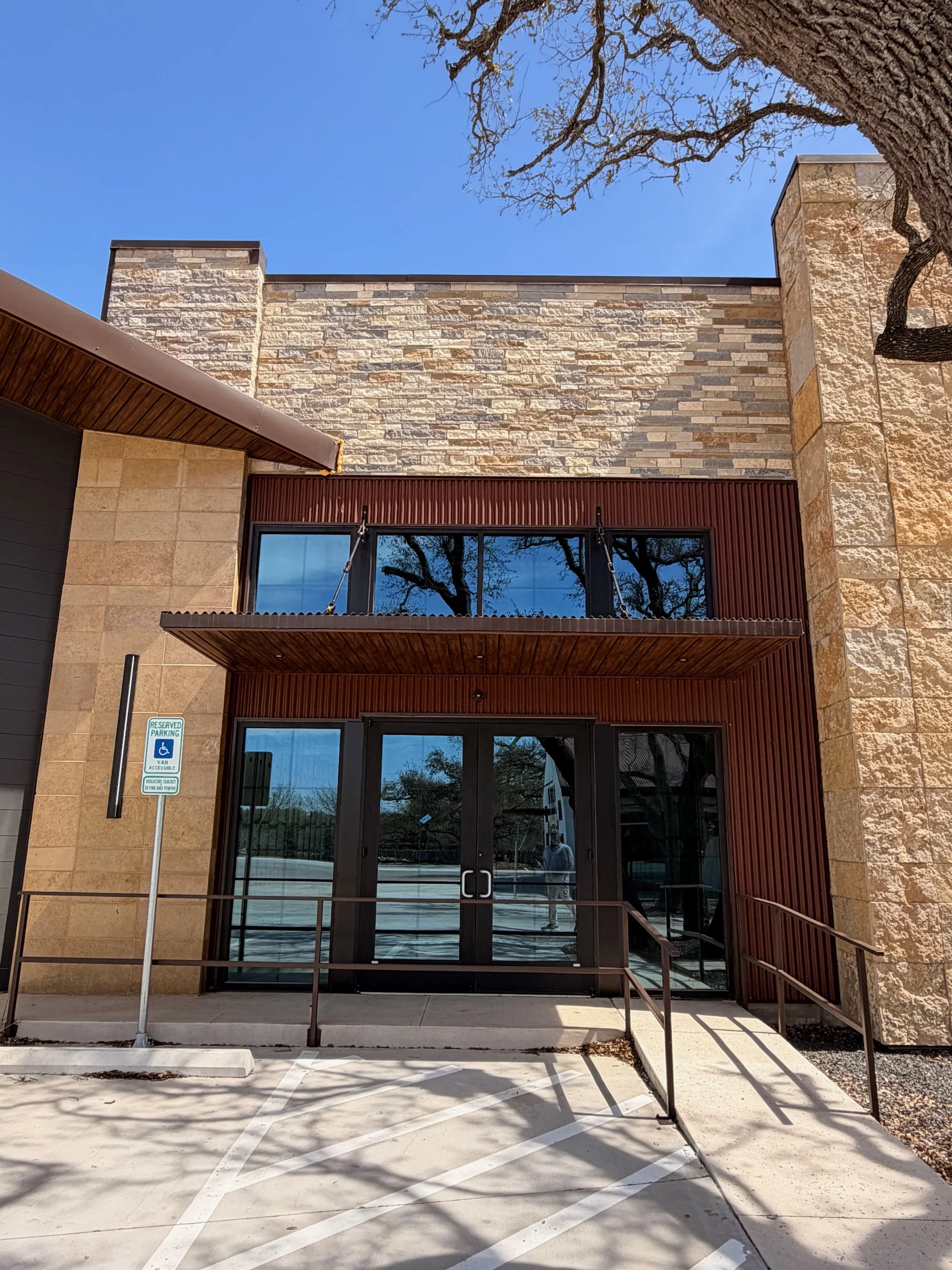 Front entrance of a modern building with glass double doors, a brown exterior metal awning, beige brick walls, and a parking space with a reserved handicap sign.