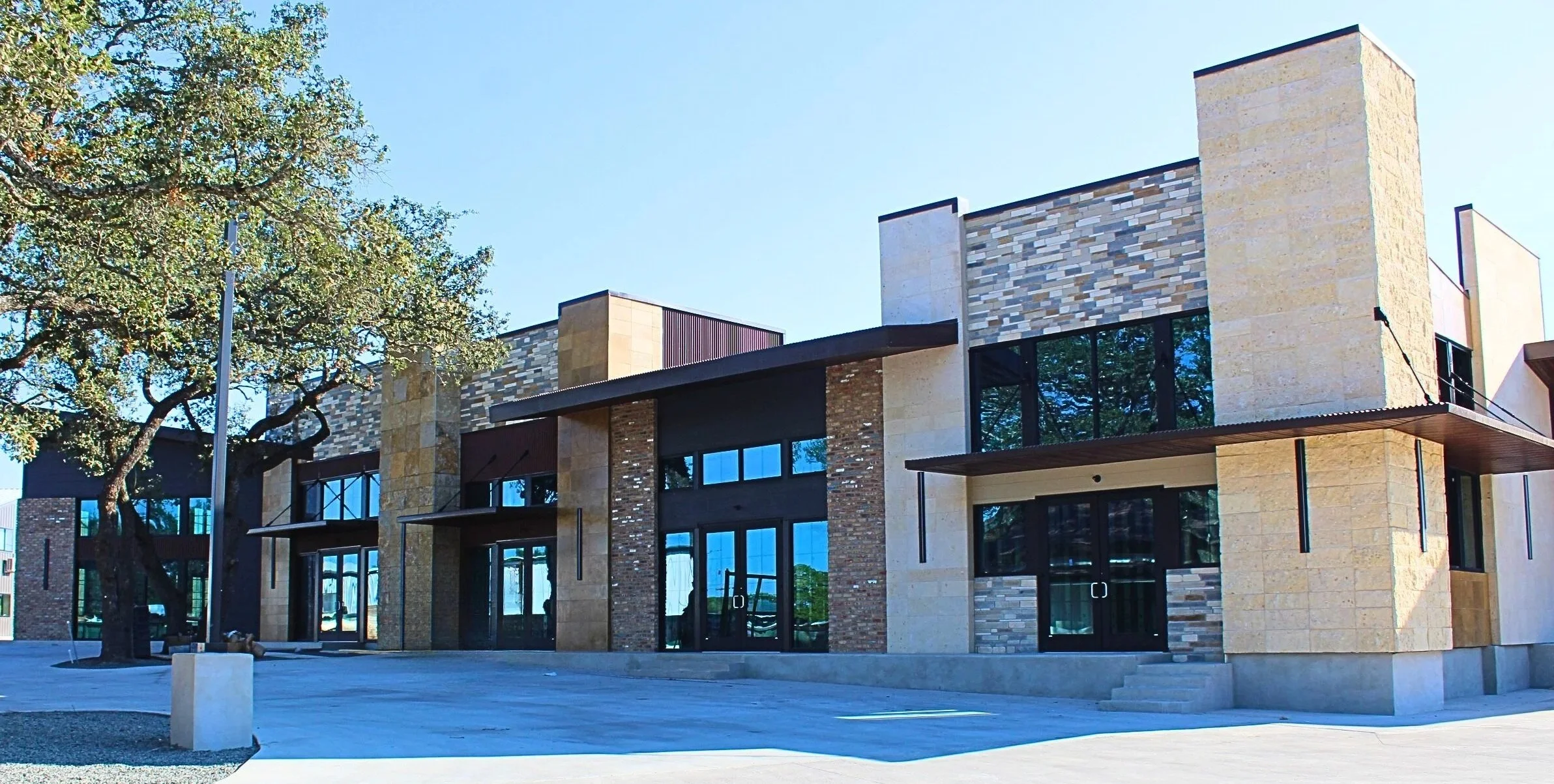 Modern multi-story commercial building with large glass windows, stone and brick facade, and a paved open area in front, under a clear blue sky.